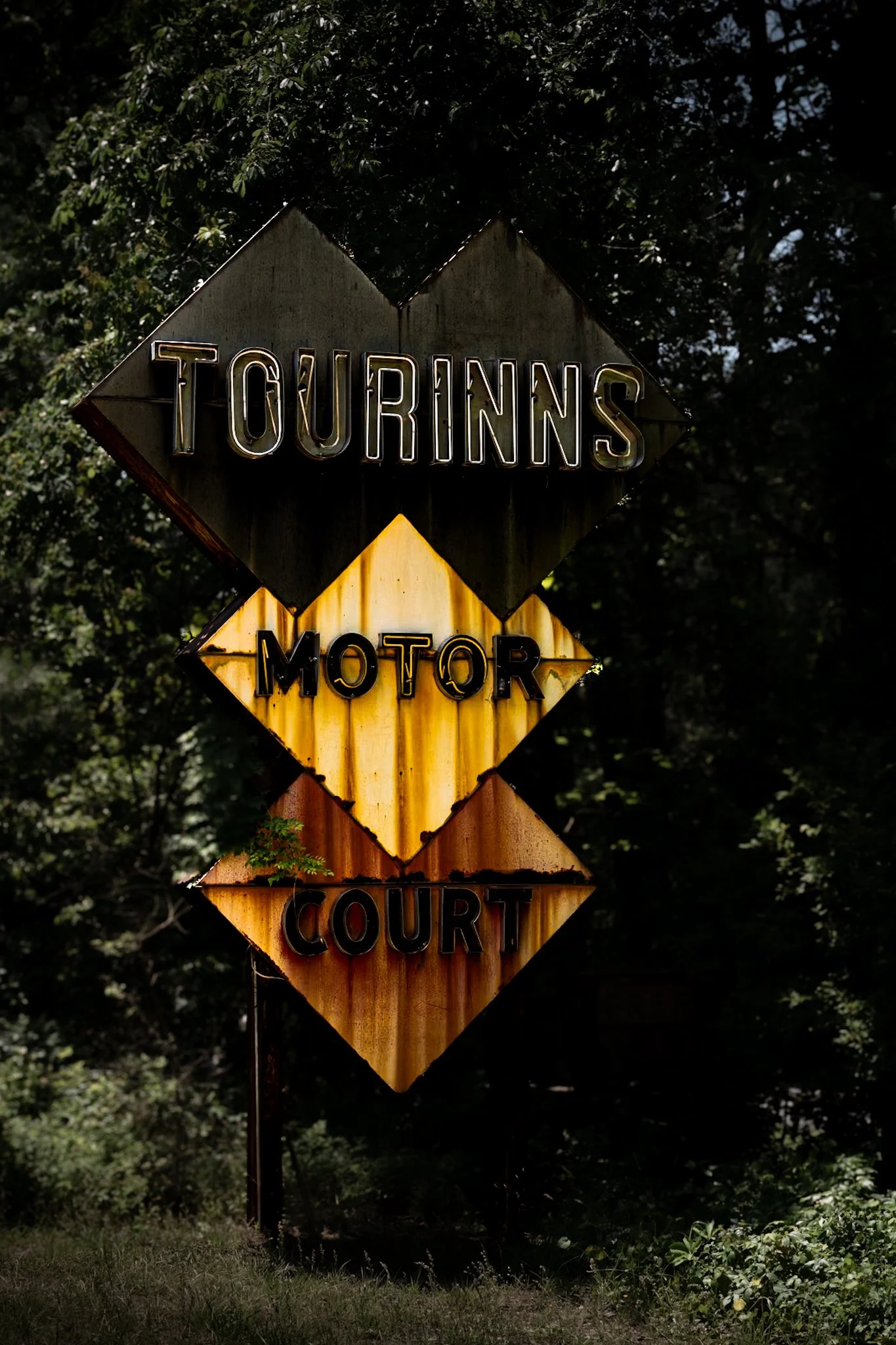 A vintage vertical sign with four diamond-shaped sections reading 'TOURINNS MOTOR COURT' in illuminated letters, surrounded by trees and foliage.