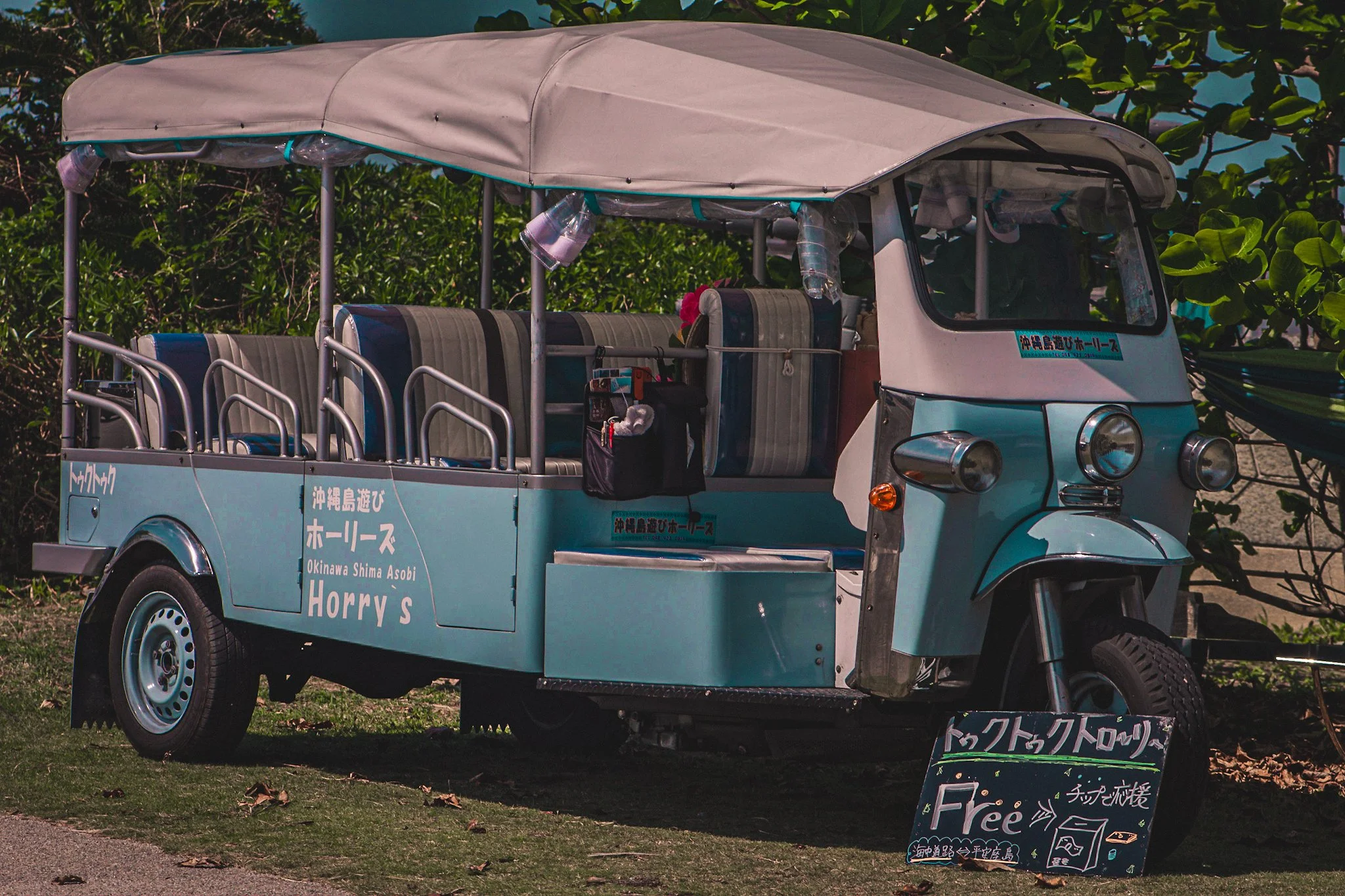 A turquoise Tuk Tuk with a beige roof parked in front of green bushes, with signage in Japanese and English advertising free services at Okinawa Shima Asobi in Japan.
