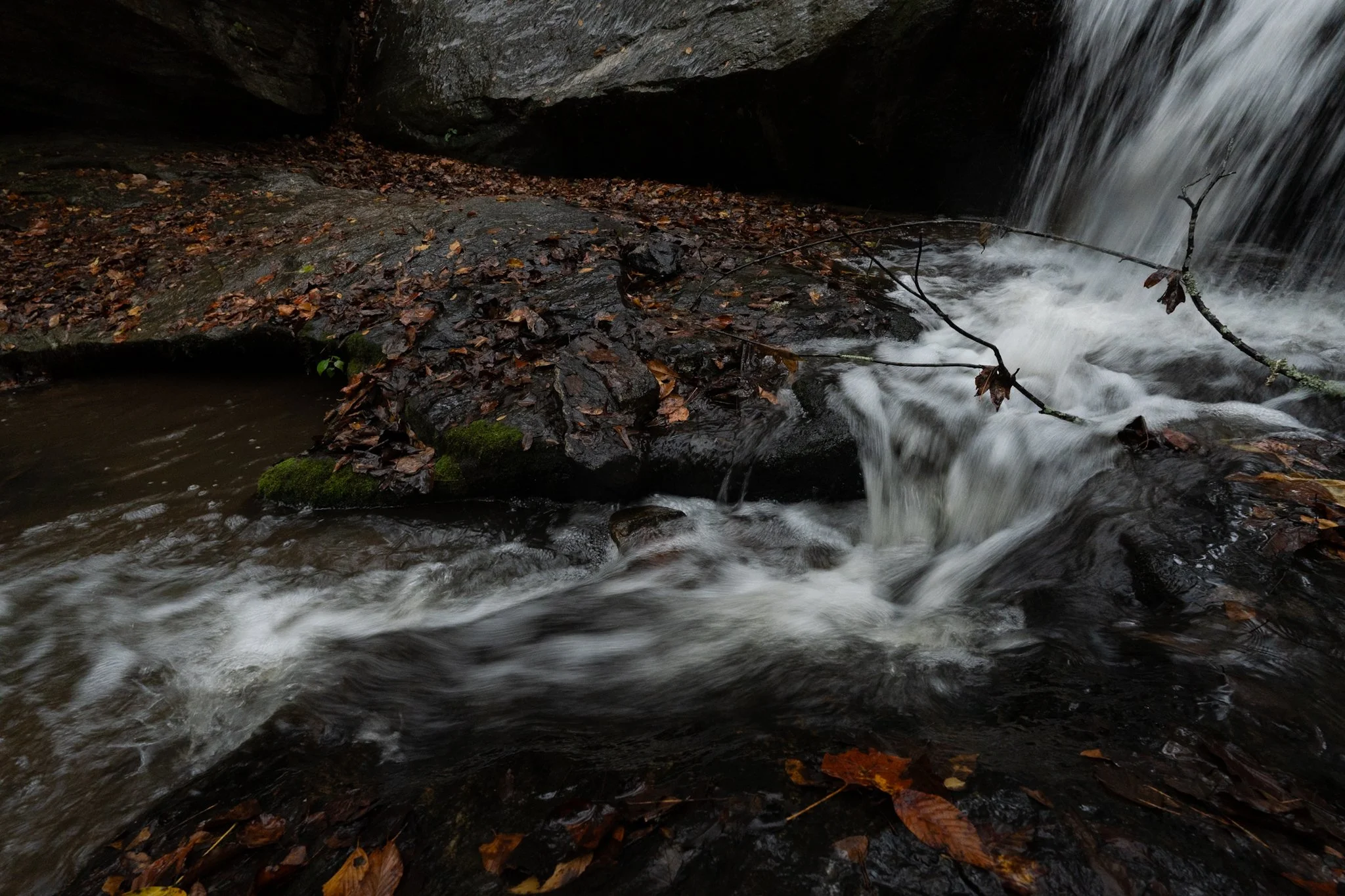 A small waterfall flowing over rocks surrounded by fallen autumn leaves in a forest.