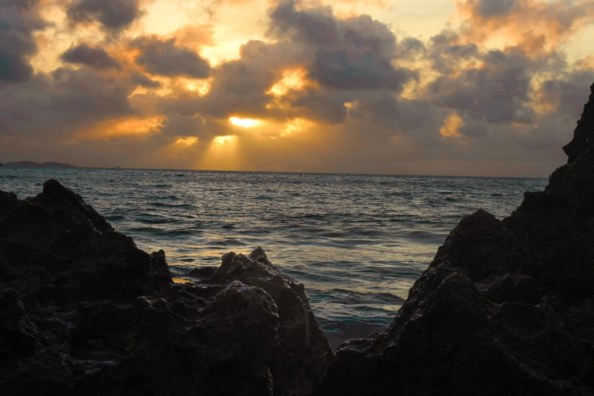 Sunset over the ocean with partially cloudy sky, viewed from a rocky shoreline.