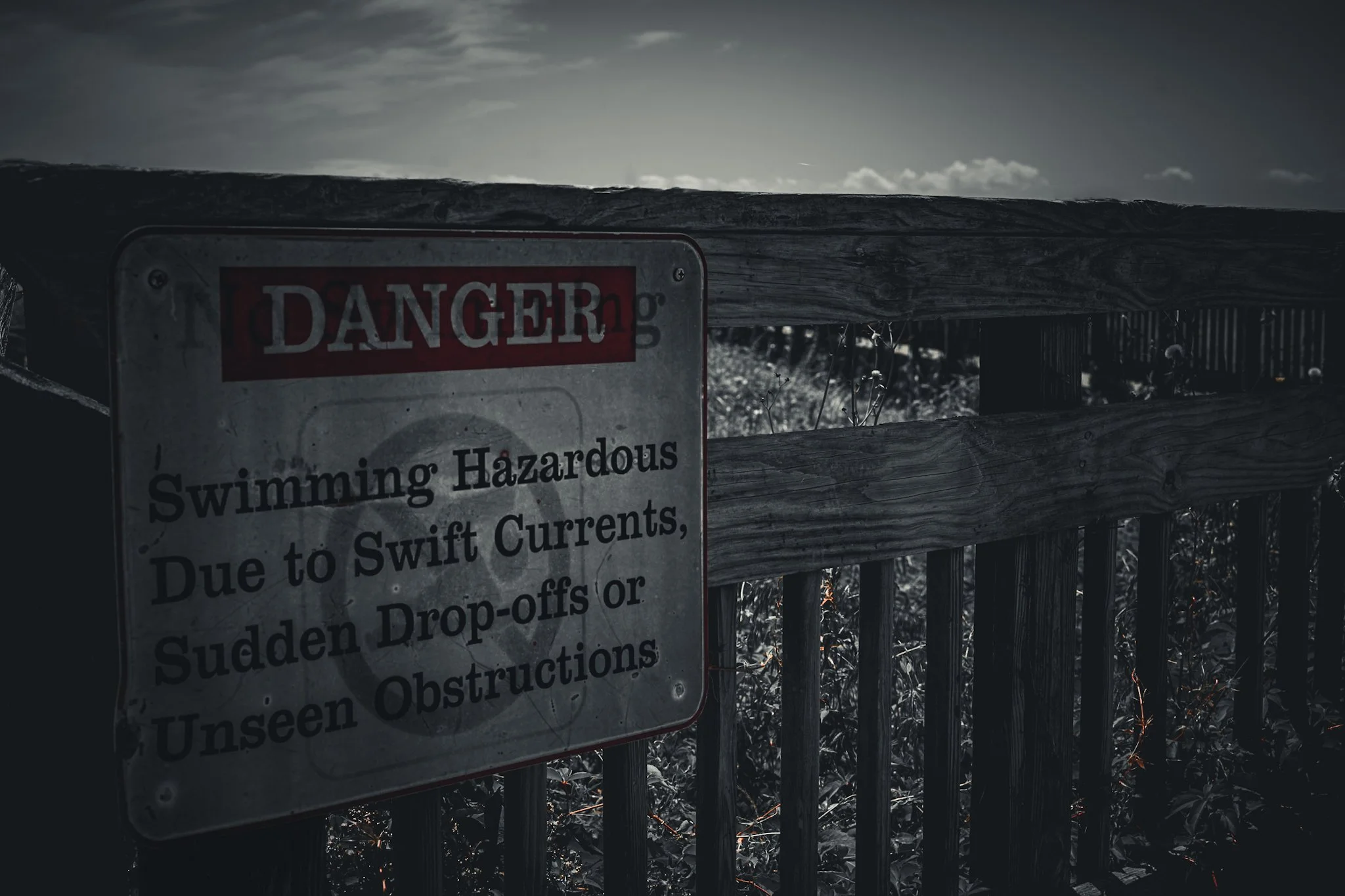 A weathered warning sign on a wooden fence warns of swimming hazards and dangerous currents, partially obscured by shadows and overgrowth.