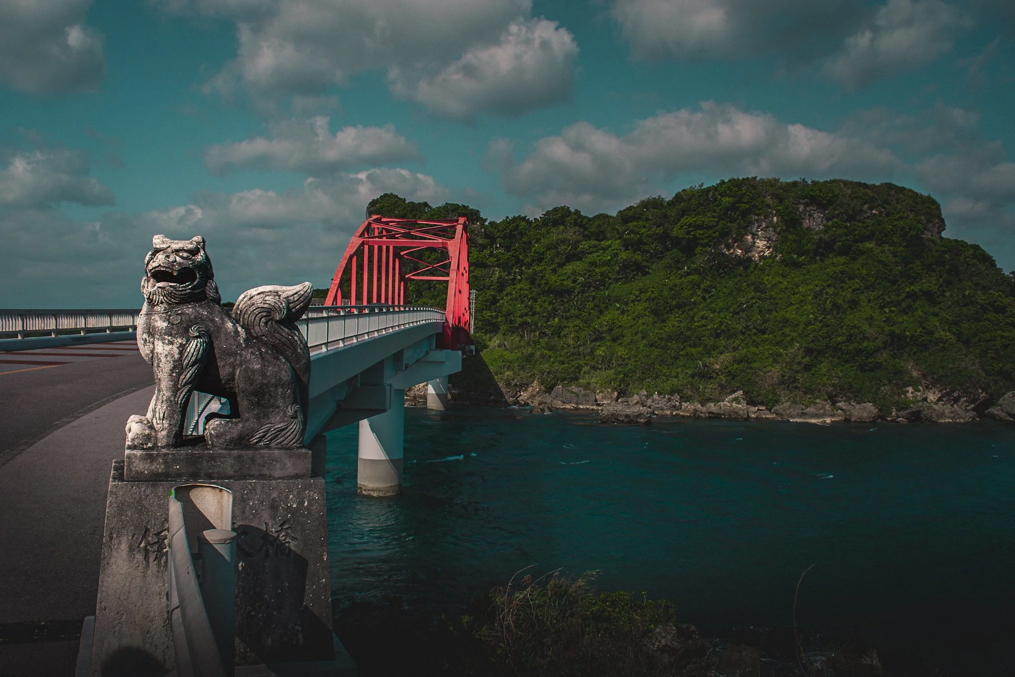 A stone statue of a mythical lion-like creature on a bridge railing, a red bridge crossing over water, with a forested island and cloudy sky in the background.