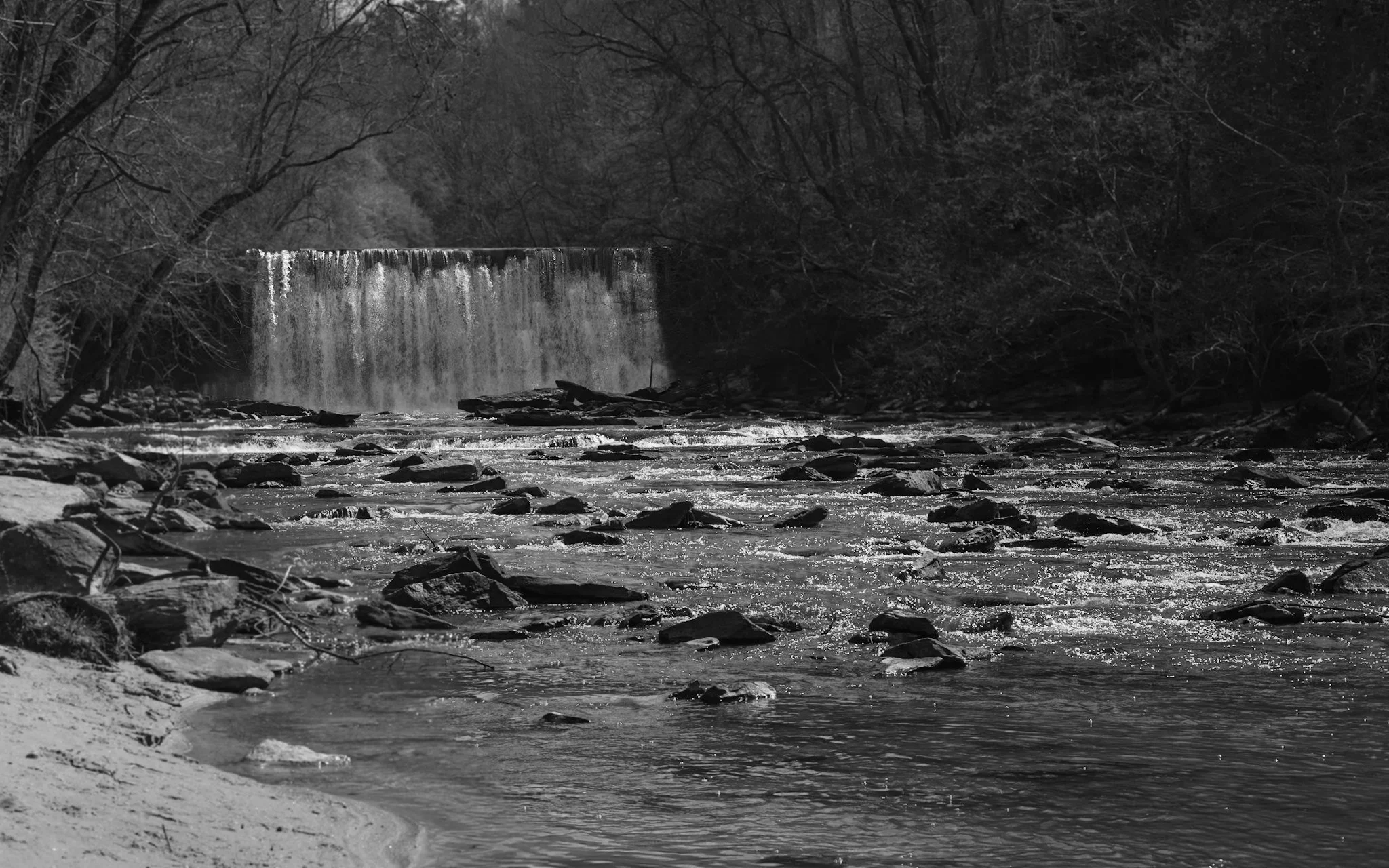 A black and white photo of a river with a small waterfall, surrounded by rocks and trees.