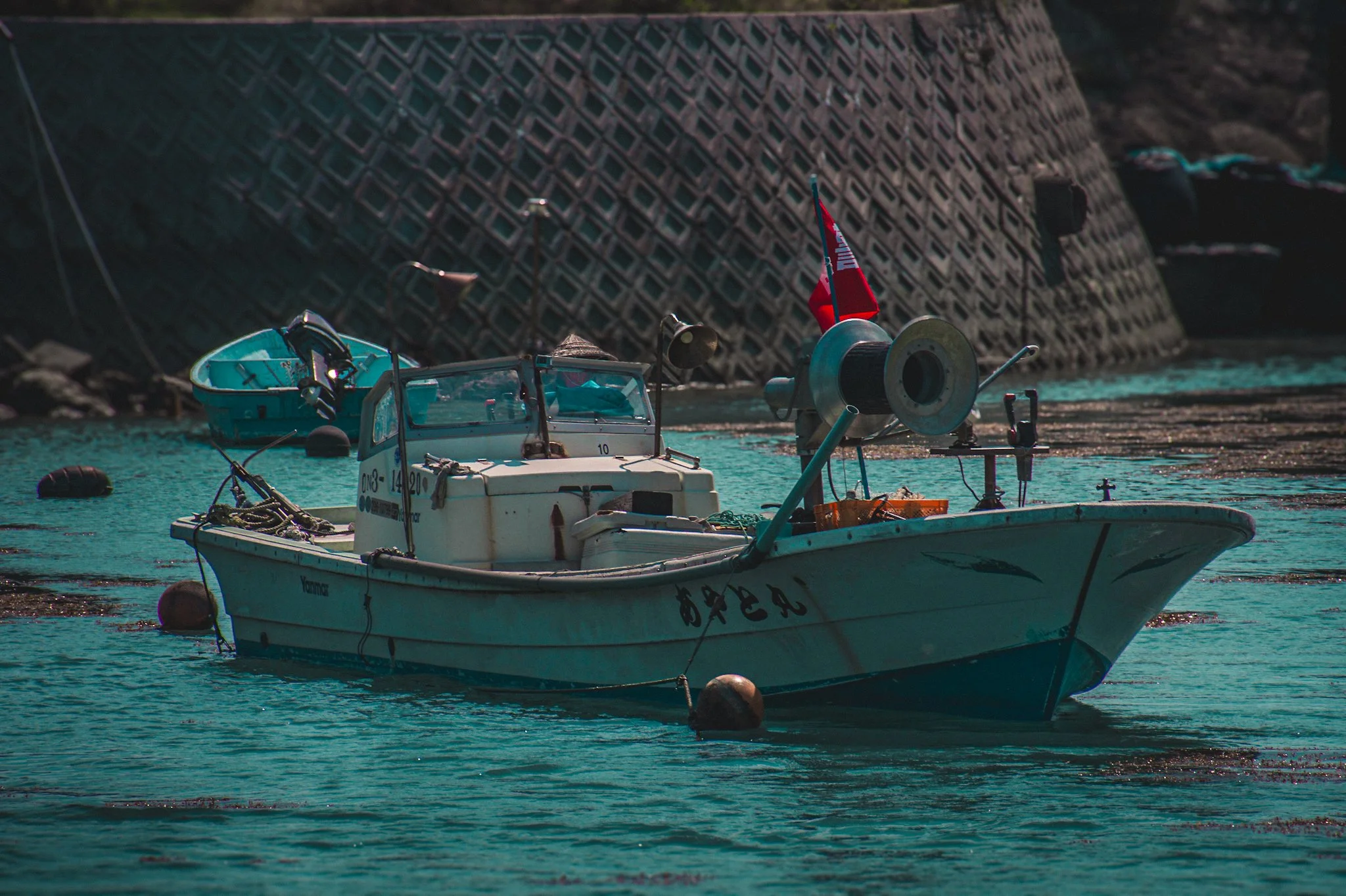 A small white boat docked in a harbor next to a large concrete seawall with a red flag attached, with a turquoise water background.