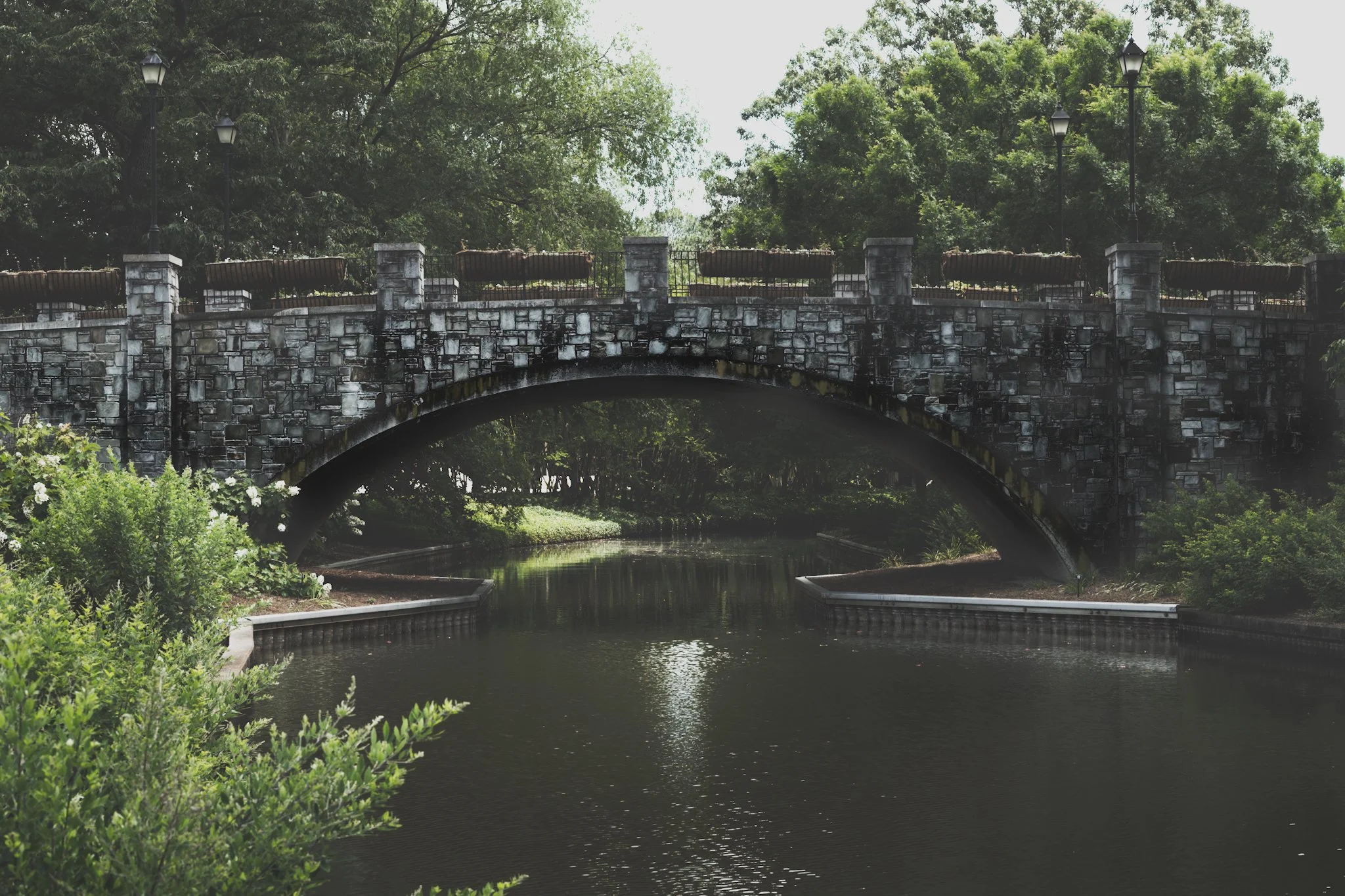 A stone bridge over a canal surrounded by green trees and bushes, with street lamps on top of the bridge.