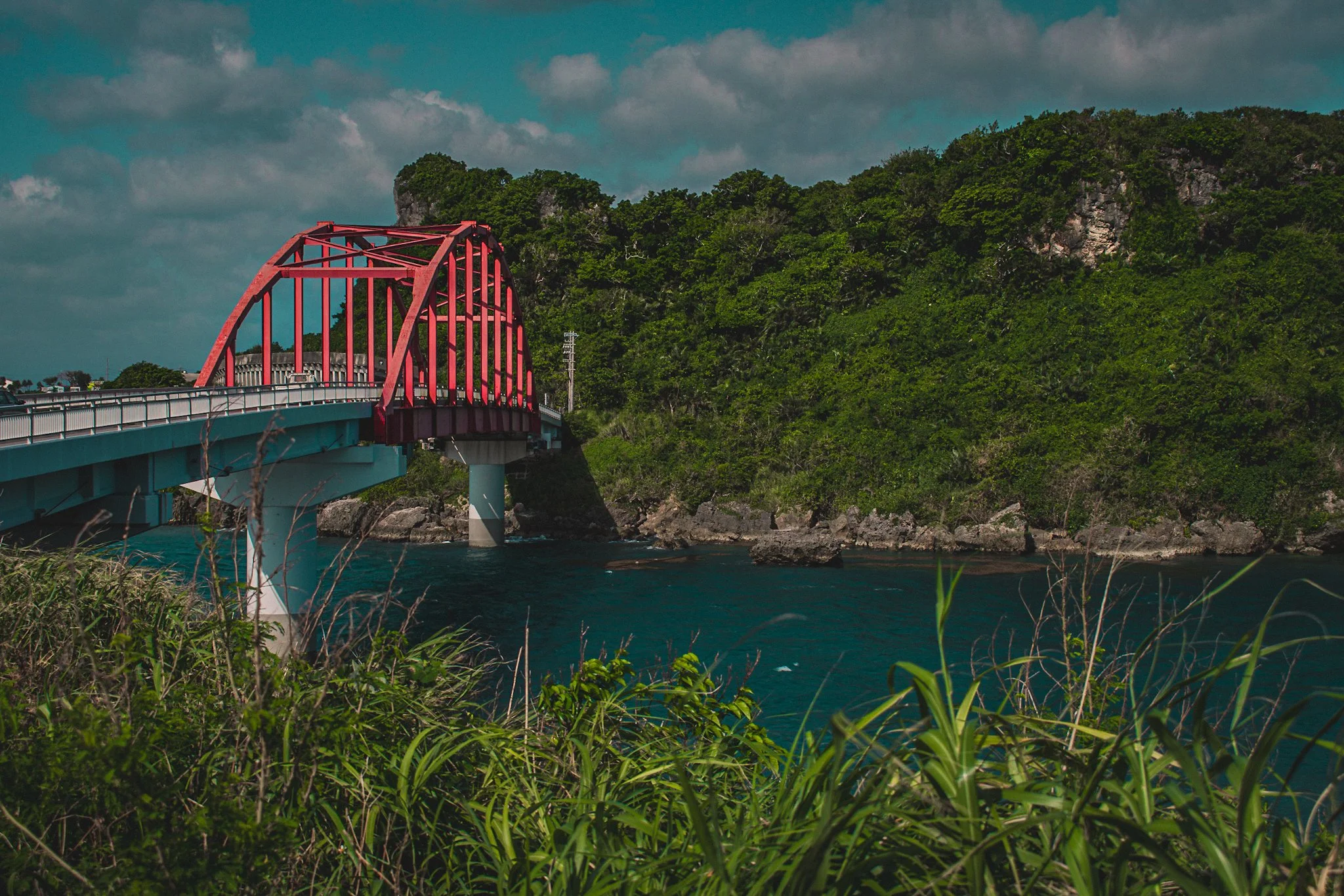 Red arch bridge over a river with green trees and rocky hills in the background under cloudy sky.