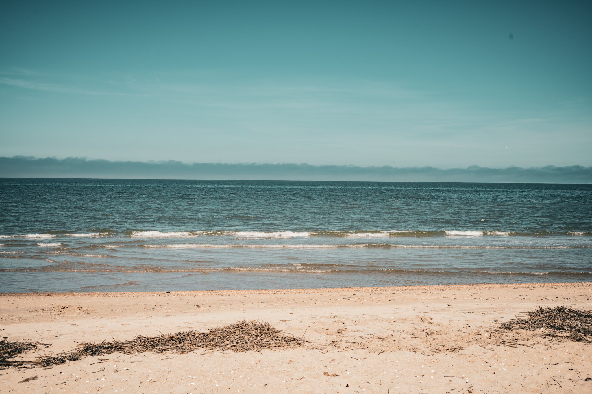 View of the ocean with gentle waves, sandy beach in the foreground, and a mostly clear sky with some clouds on the horizon.