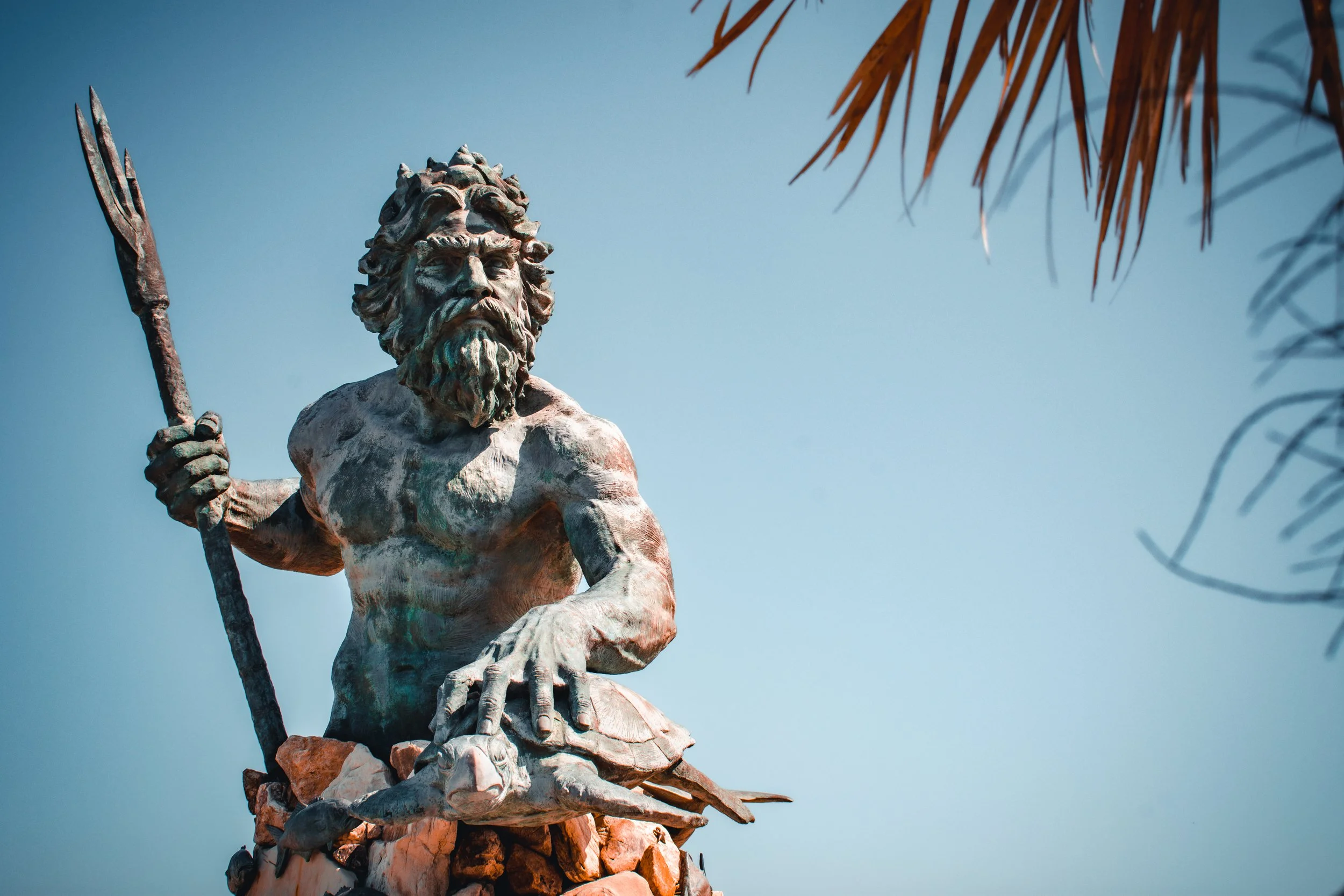 Statue of a bearded man holding a trident, sitting on rocks, with palm leaves and a blue sky in the background.