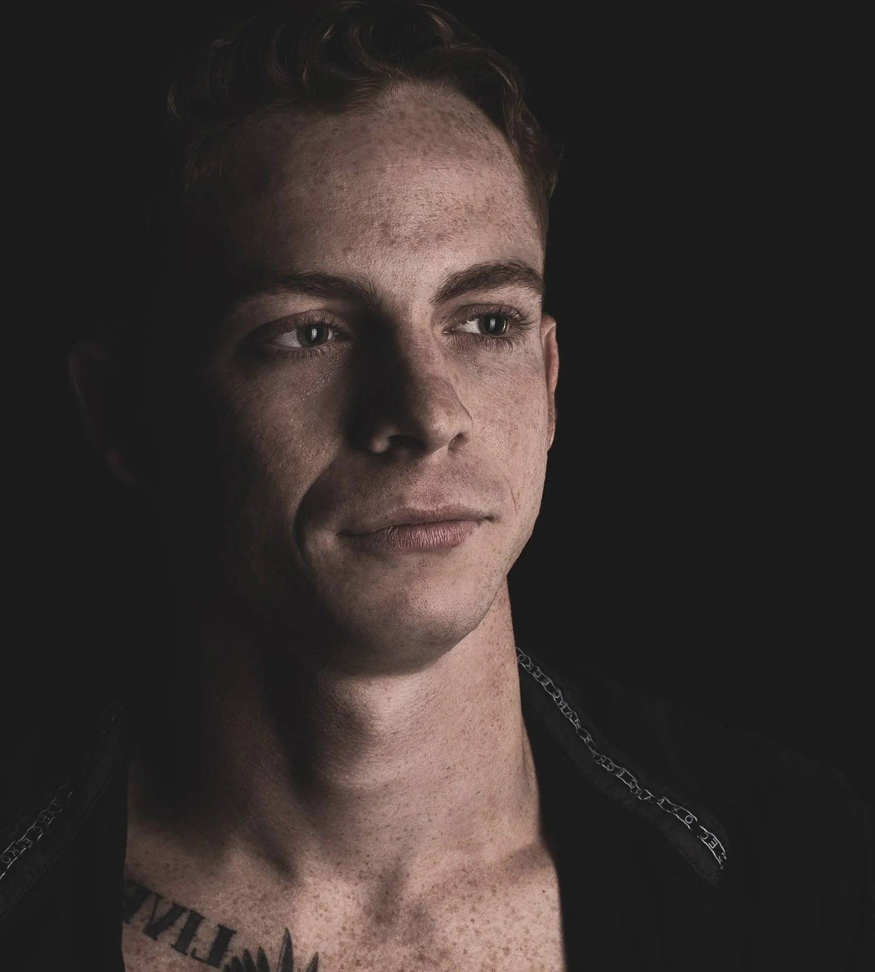 Close-up of a young man's face with short brown hair, freckled skin, and light-colored eyes, set against a dark background.