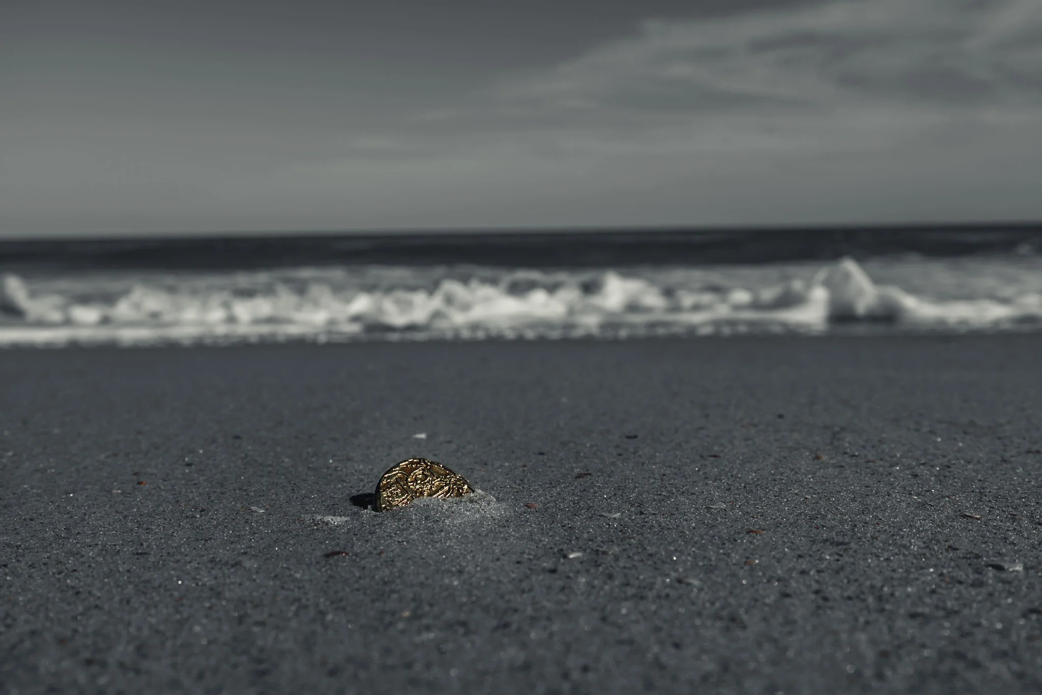 A shiny gold coin partially buried in the dark sandy beach with ocean waves in the background under a gray cloudy sky.