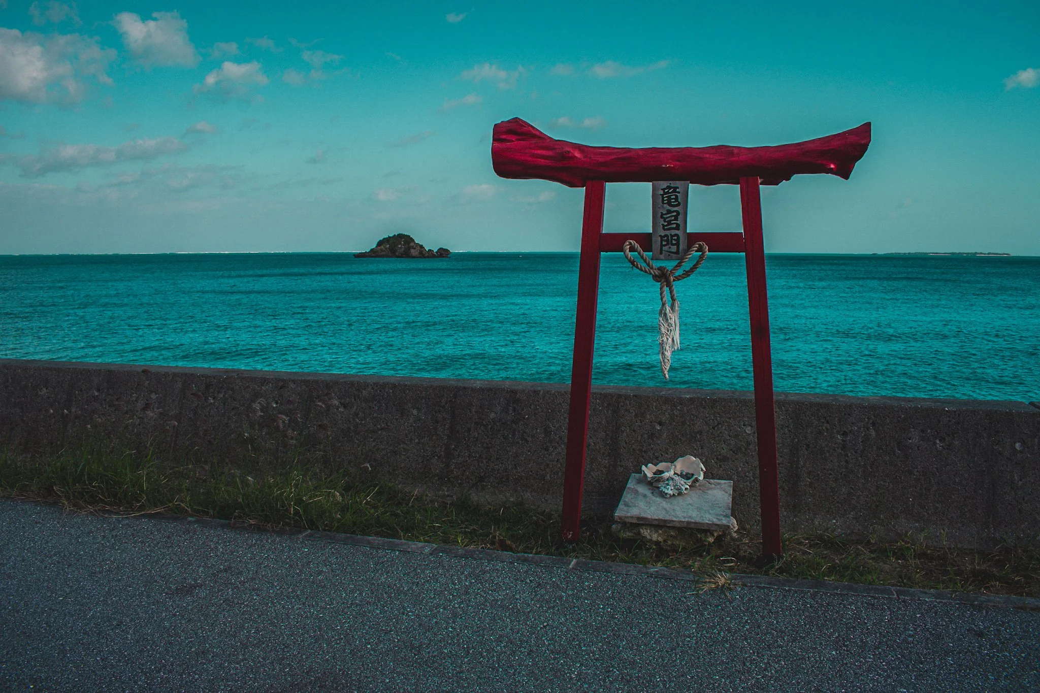 A red torii gate by the ocean, with a small island in the distance and a partly cloudy sky.