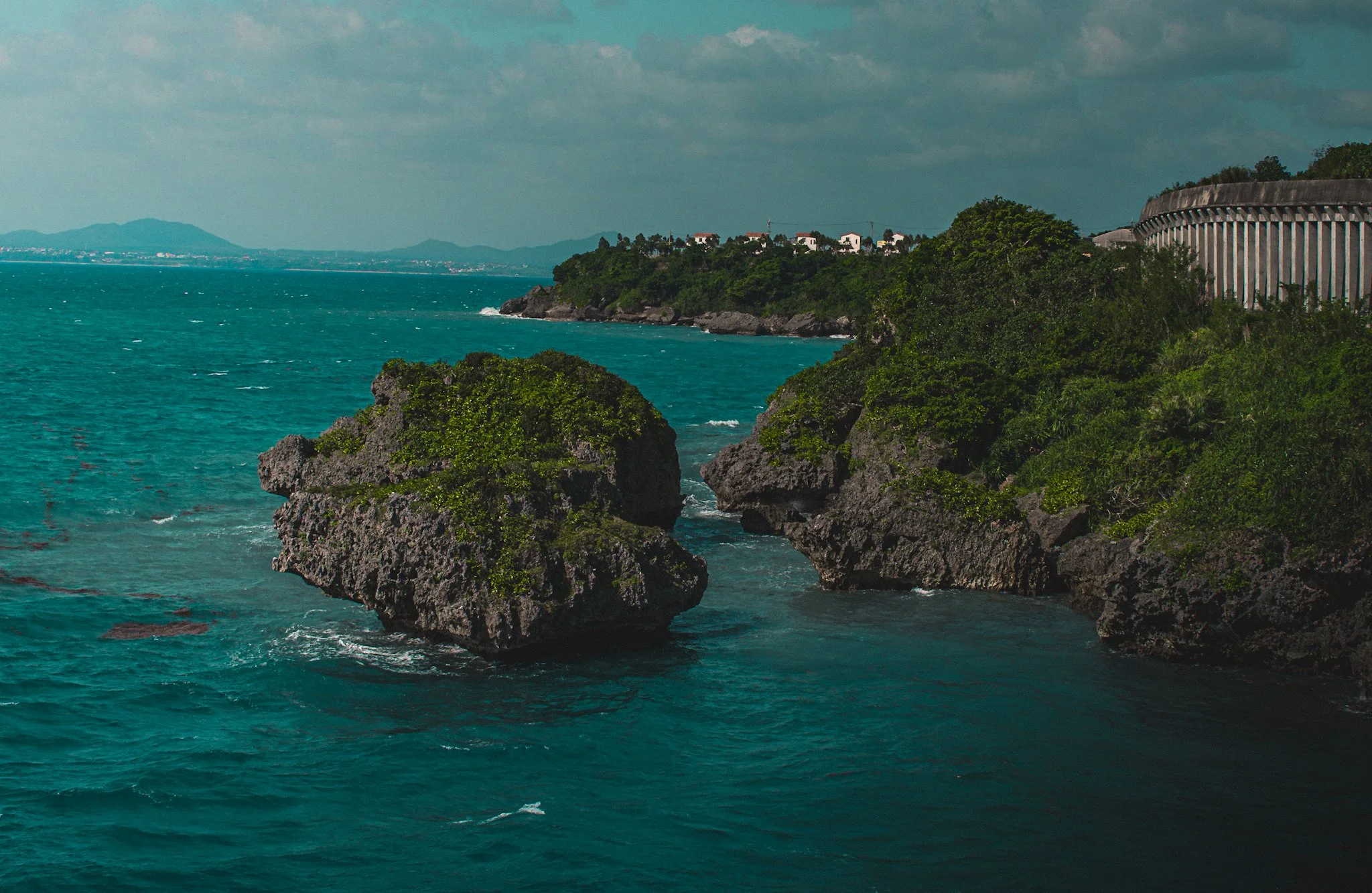 Seascape with turquoise water, rocky cliffs with greenery, and a distant shoreline with buildings and hills under a cloudy sky.