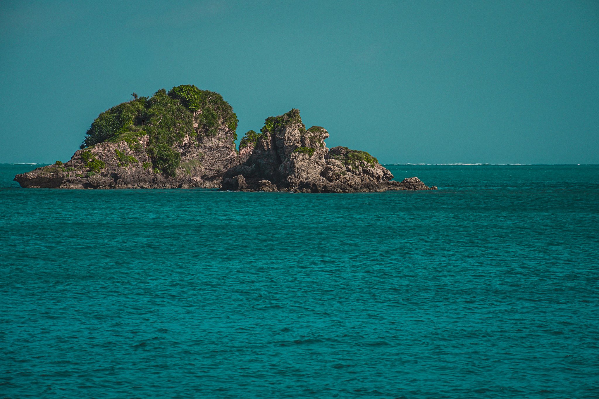 A small rocky island with green vegetation surrounded by turquoise water under a clear blue sky.
