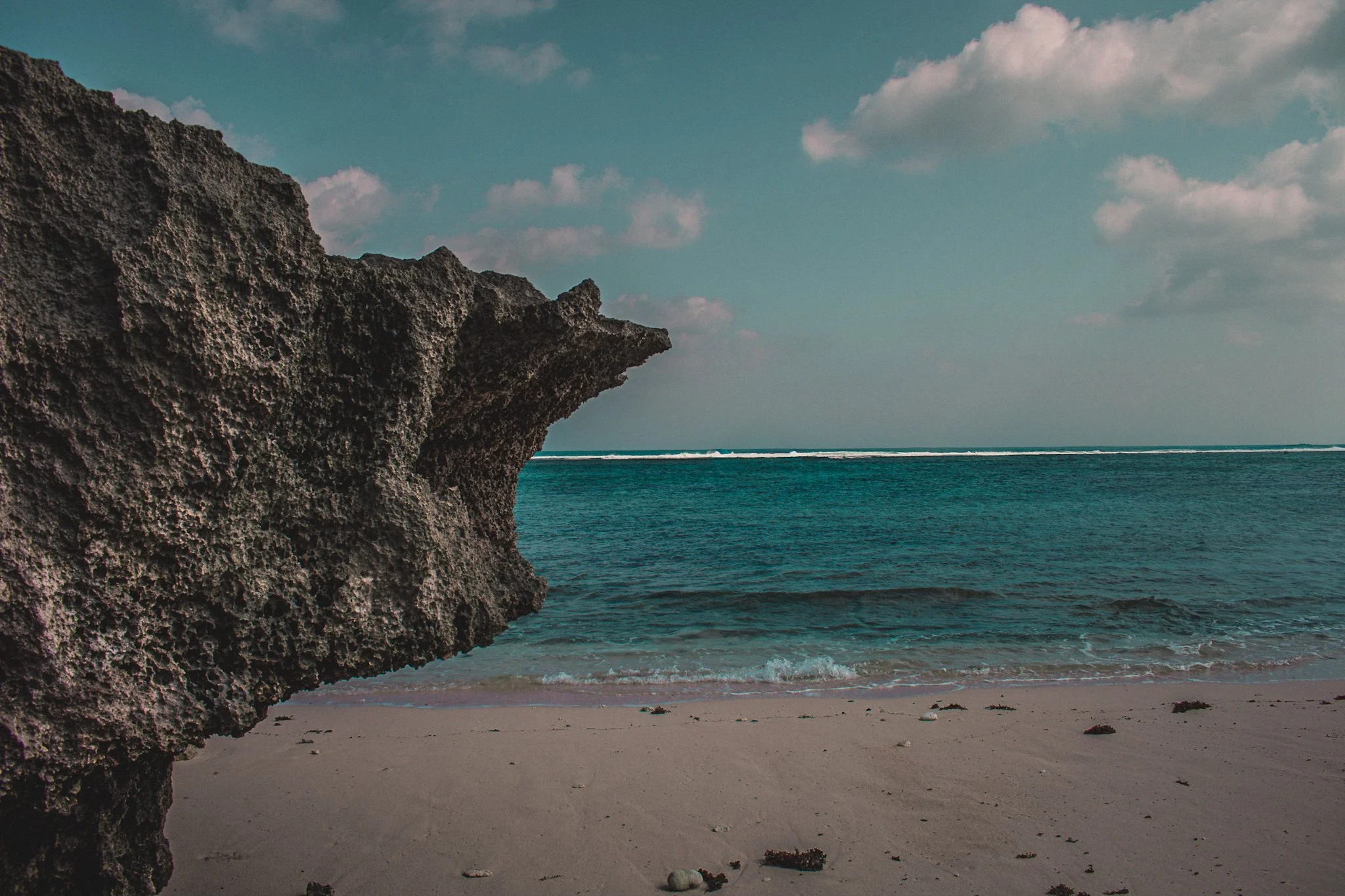 A rocky cliff on the left overlooking a sandy beach and the ocean under a partly cloudy sky.