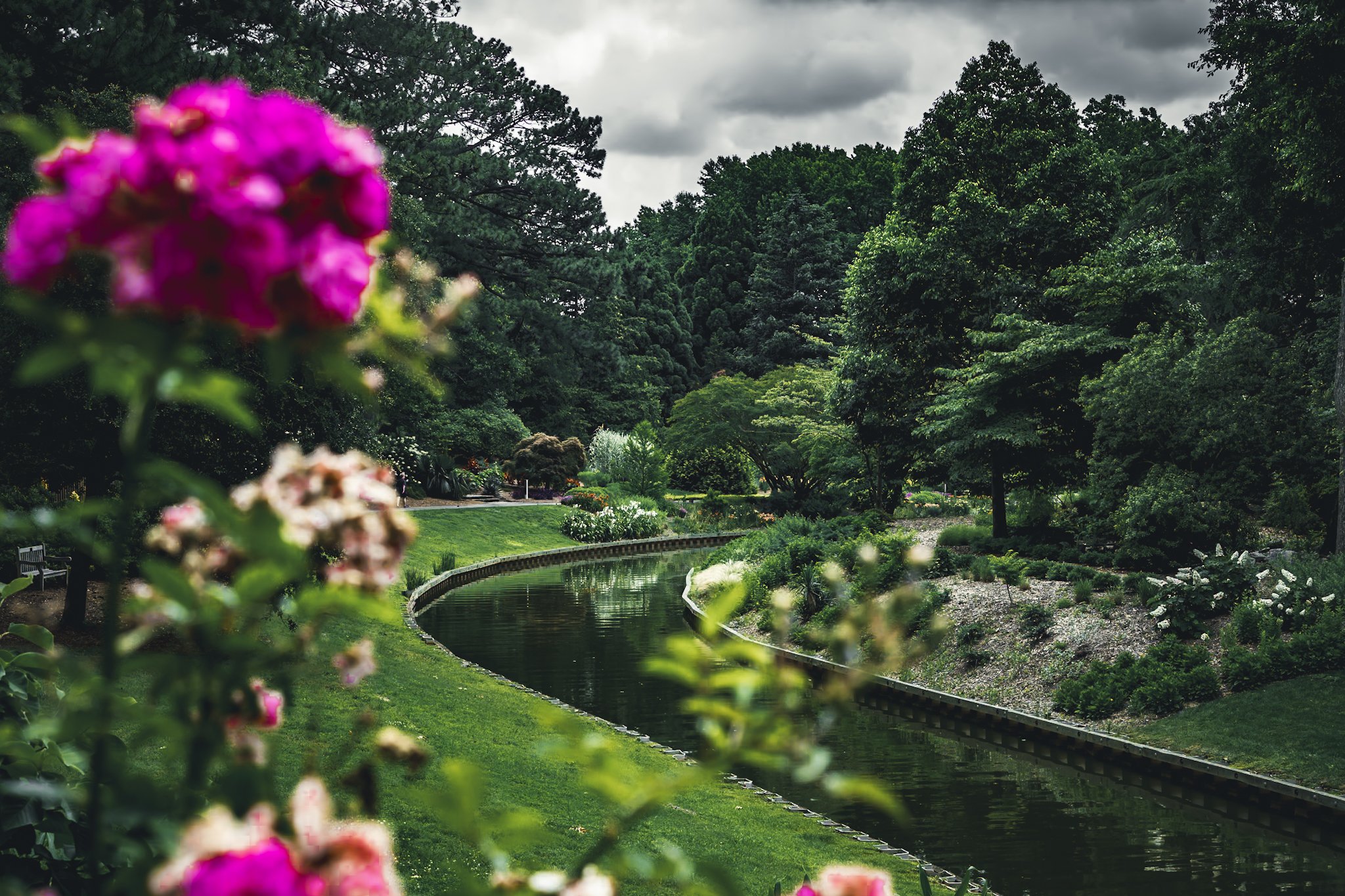 A peaceful park with a curved waterway, lush green trees, and blooming flowers on a cloudy day.