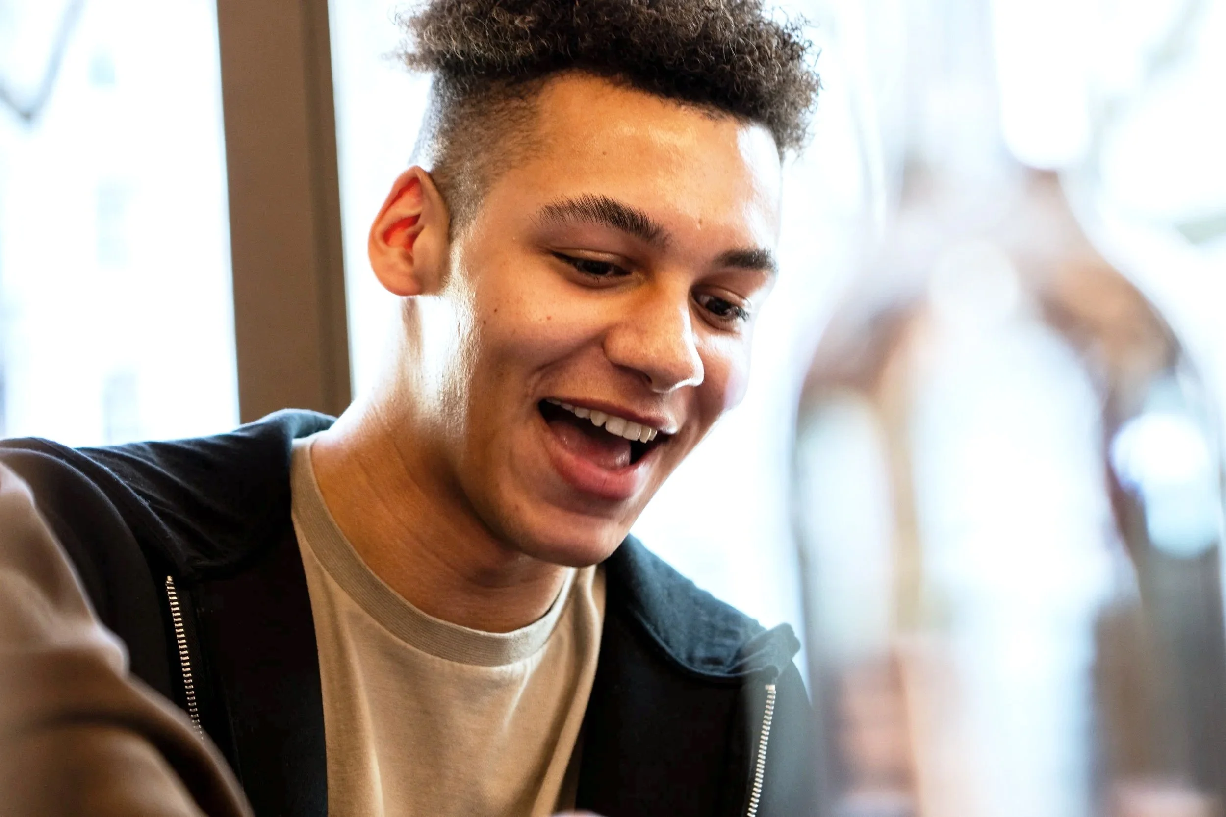 A young man with curly hair, smiling and looking down, might be sitting indoors near a window with natural light