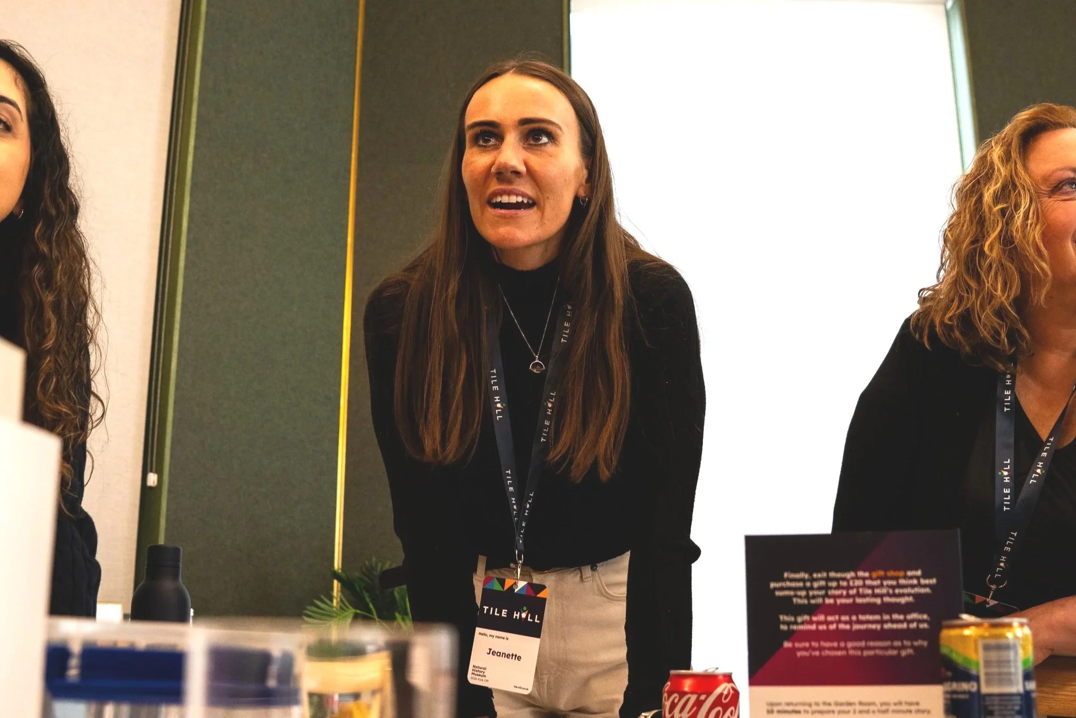Woman with long brown hair, wearing a black top and necklace, speaking at an event with a name tag that reads "Jeannette." She is standing behind a table with various items, including a Coca-Cola can and a black bottle. There are other women partially visible on either side of her, and a large screen or whiteboard behind her.