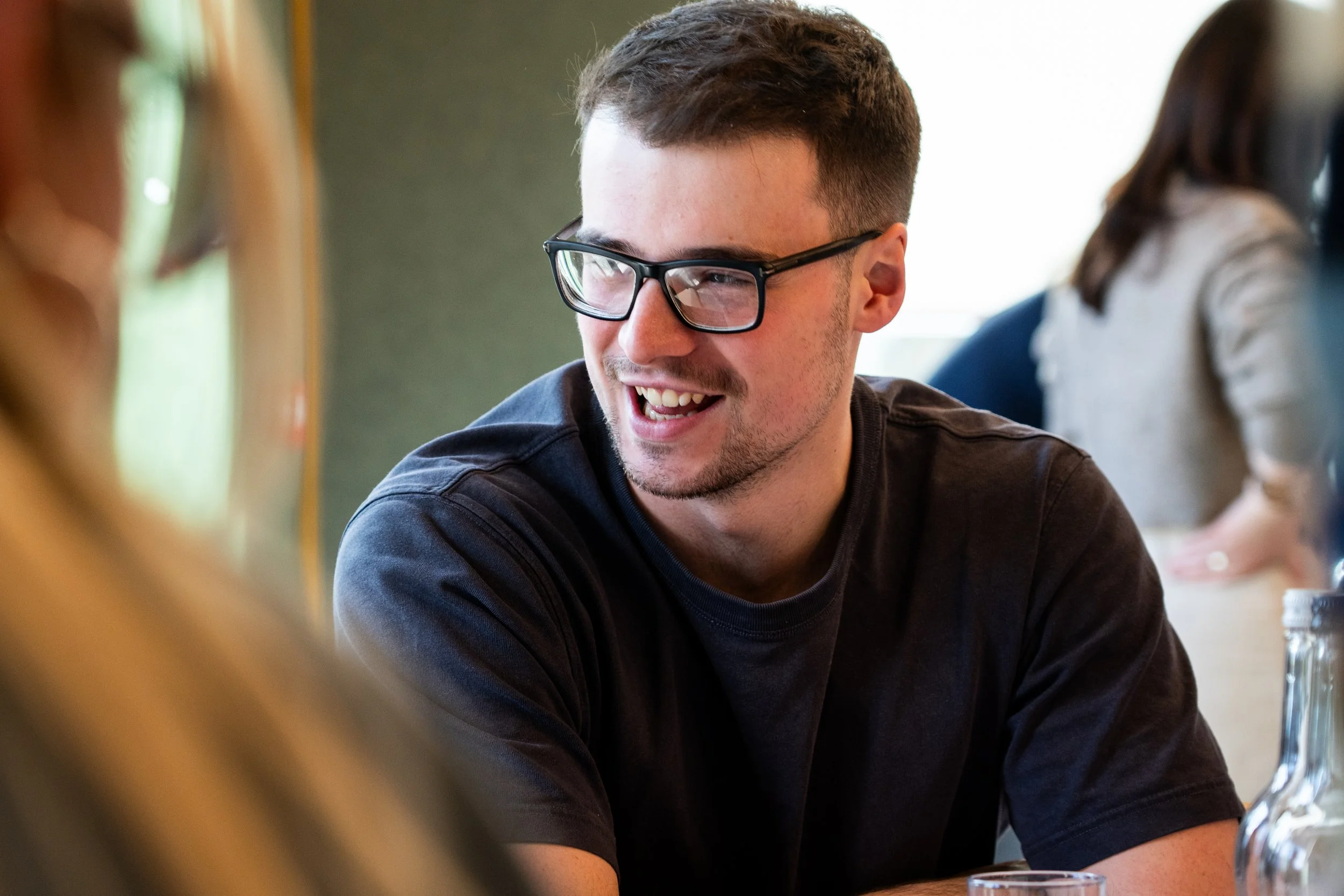 A young man with short dark hair and glasses smiling during a social gathering, sitting at a table with a drink in front of him, engaging in conversation.