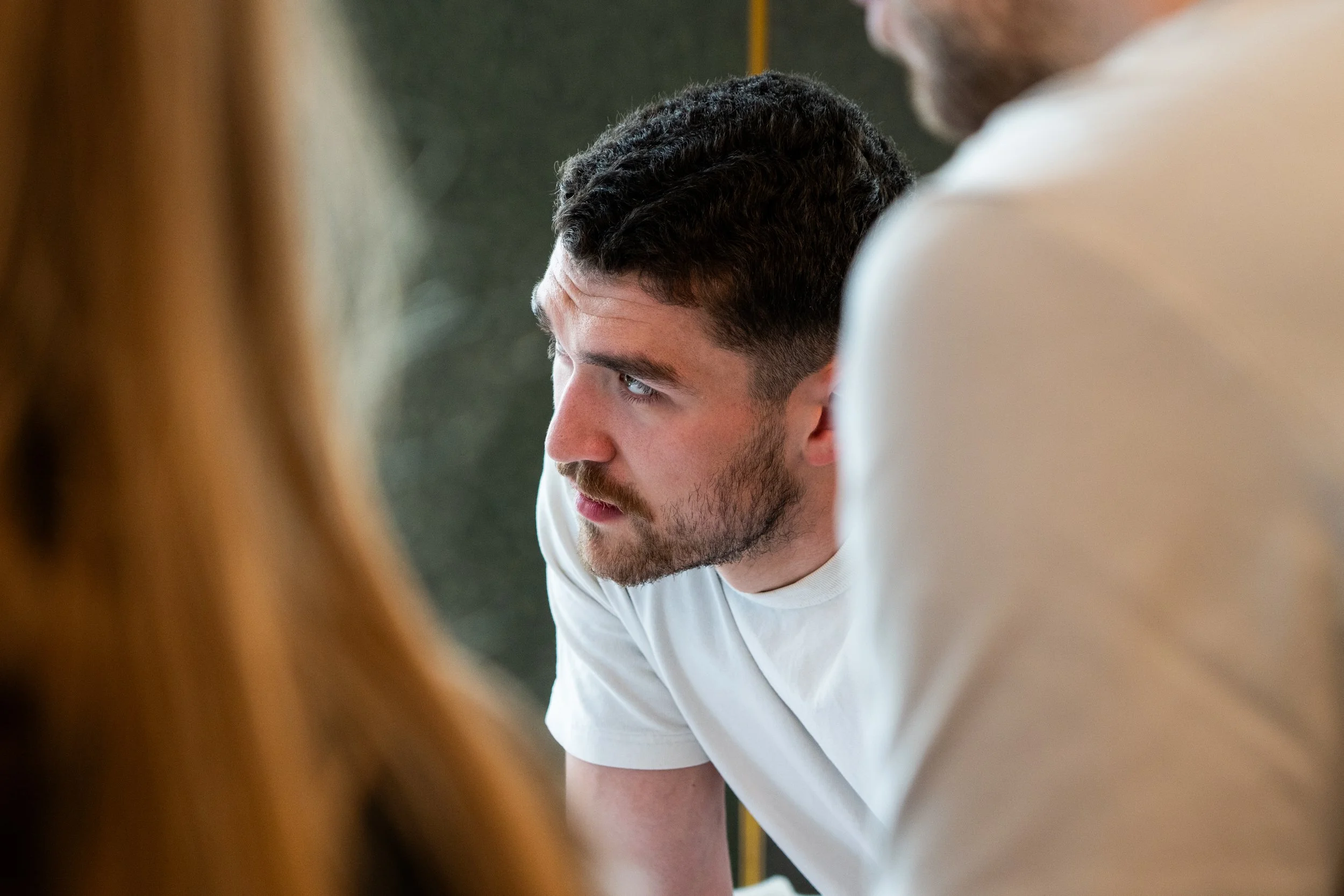 Close-up of a young man with dark, curly hair and a beard, looking to the side, framed by blurred out features in the foreground.