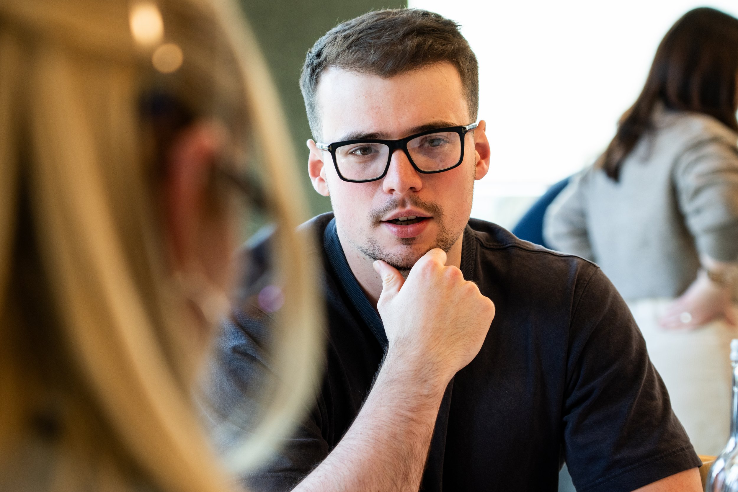 A young man with short dark hair, glasses, and a slight beard, engaged in conversation or deep thought, sitting at a table during a social or academic setting, with other people visible in the background.