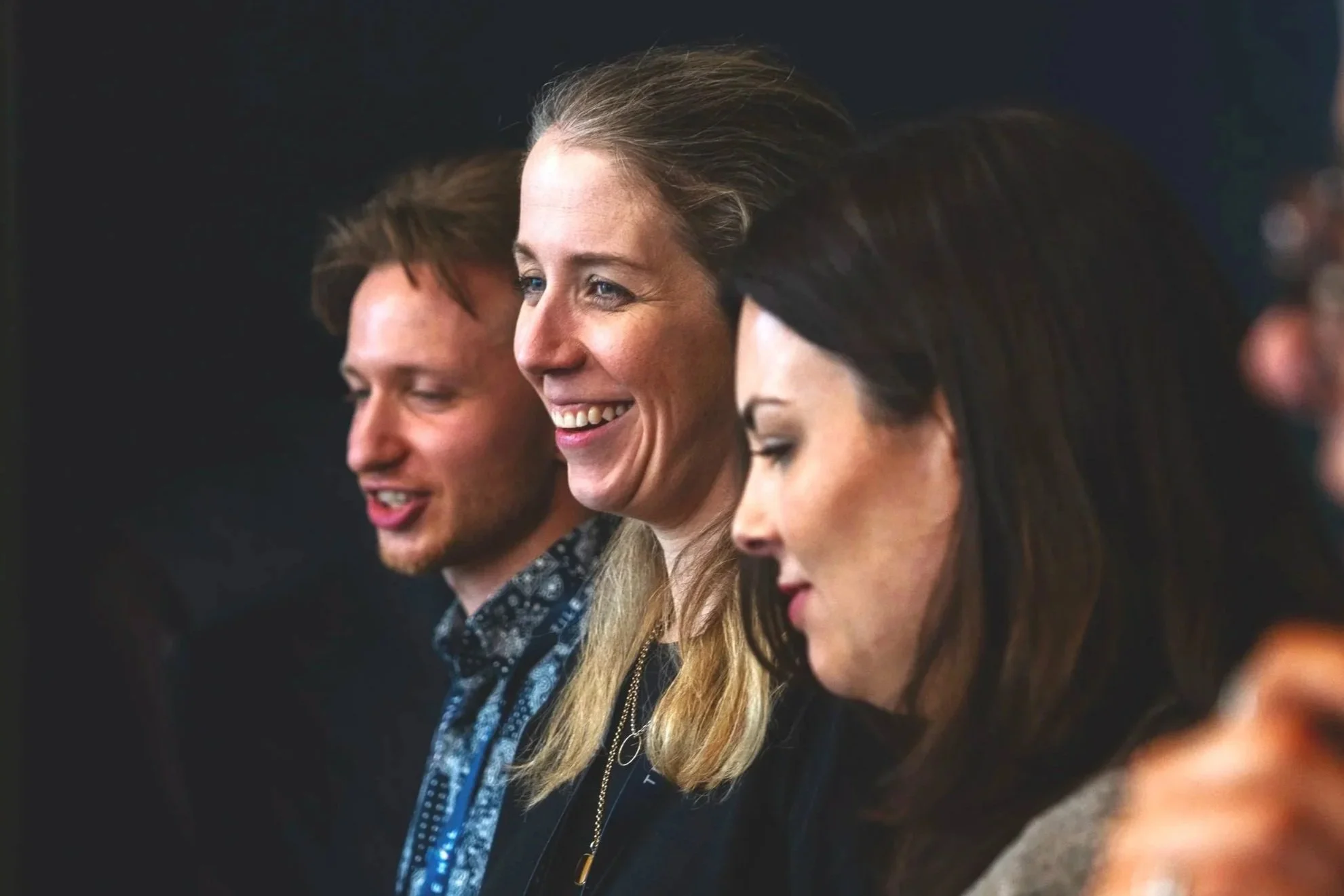 Close-up of three people with side profiles, smiling and looking down, in a dark background.