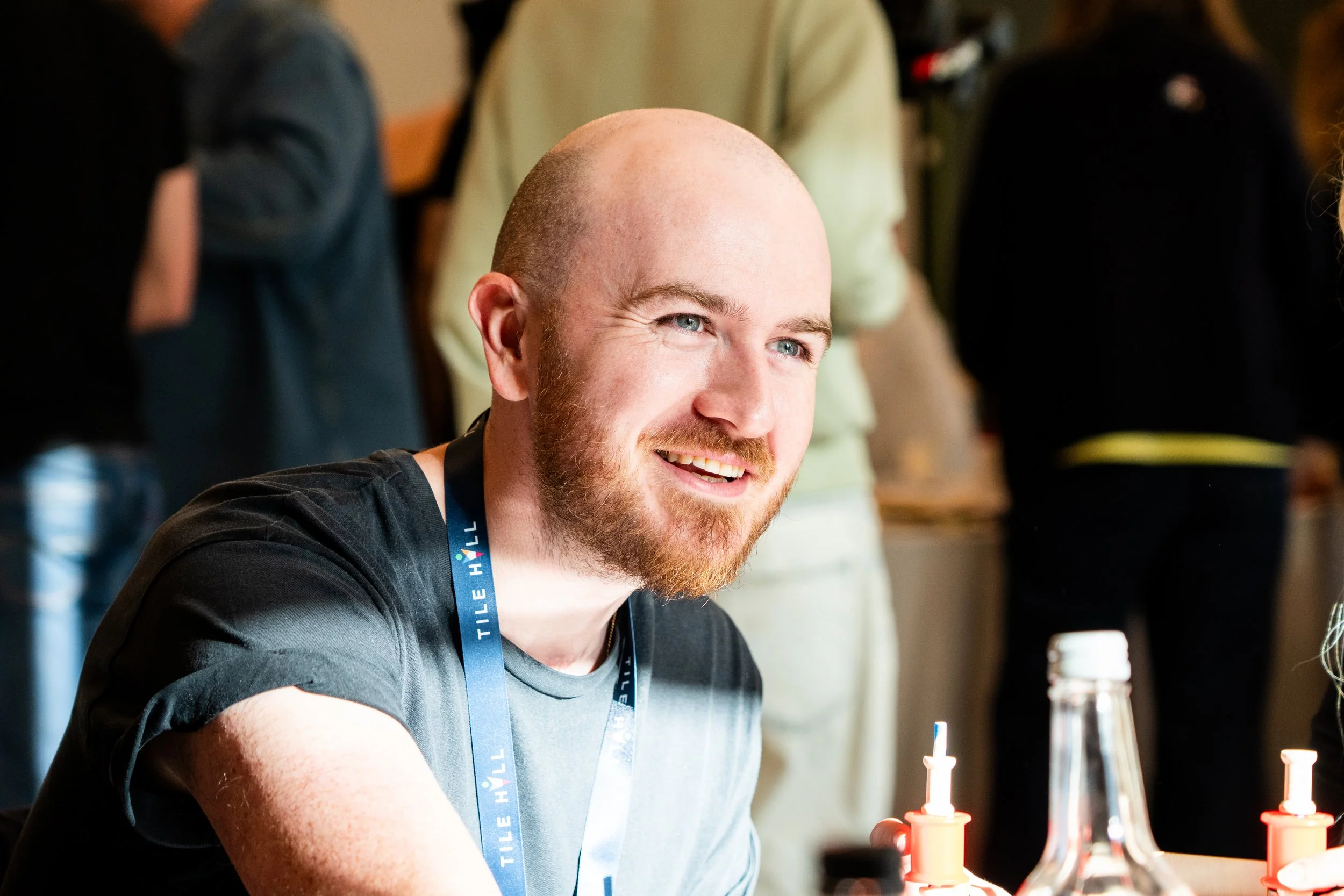 A smiling man with a beard and blue eyes sitting at a table with red and white bottles, possibly ketchup or mustard, in front of him. He is wearing a black t-shirt and has a blue lanyard around his neck. There are blurred people in the background.
