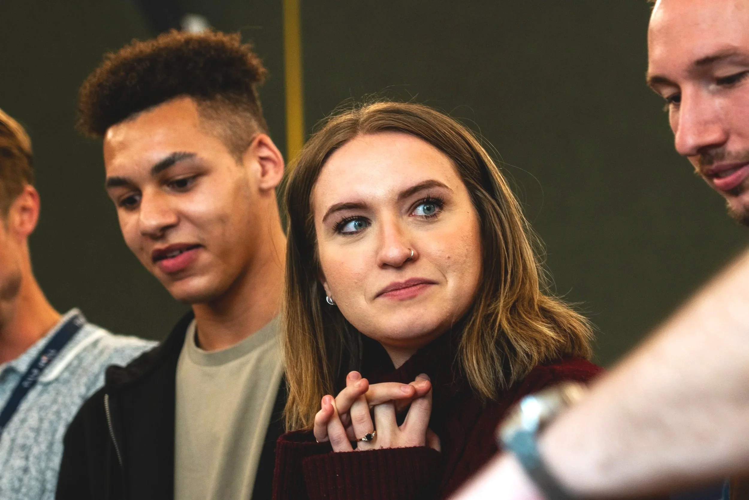 Three young adults, a woman with long brown hair and a nose ring, and two young men, stand close together indoors, appearing to be engaged in an activity or conversation.
