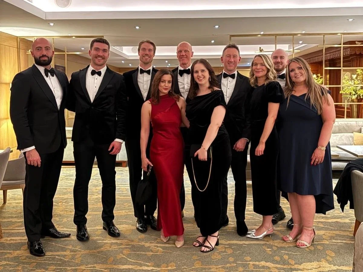 Group of ten people dressed in formal attire, including tuxedos and evening gowns, standing together in a hotel lobby or event space with warm lighting and wooden decor, smiling at the camera.