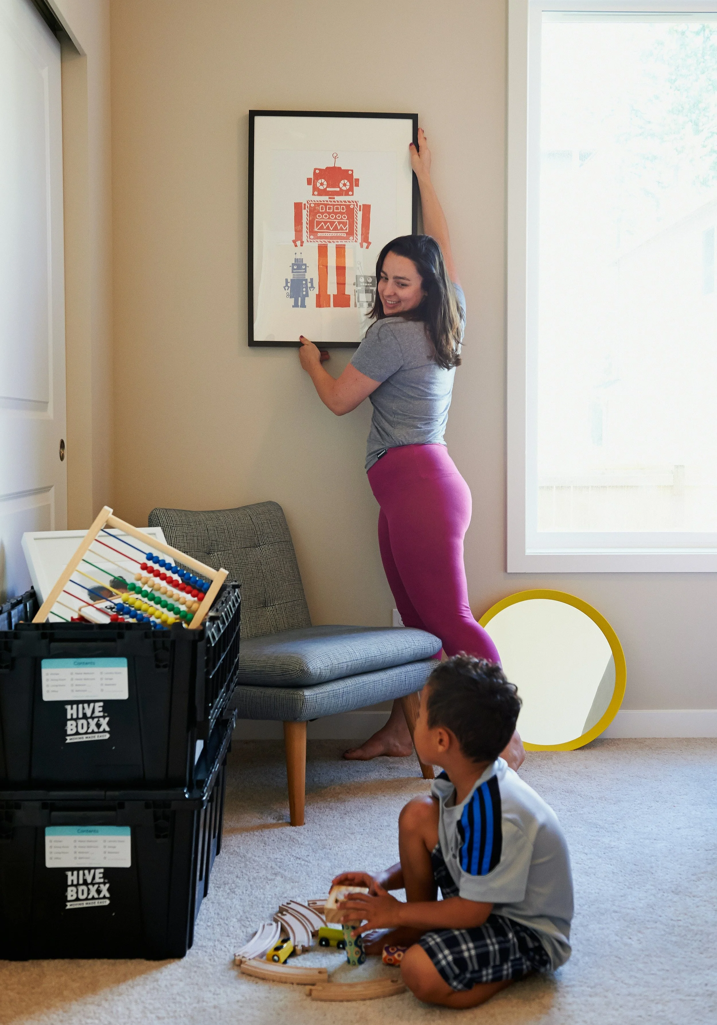 A woman is hanging up a framed artwork of a vintage-style red and blue robot in a cozy room, with children playing nearby on the carpet, toys and a wooden train set on the floor.
