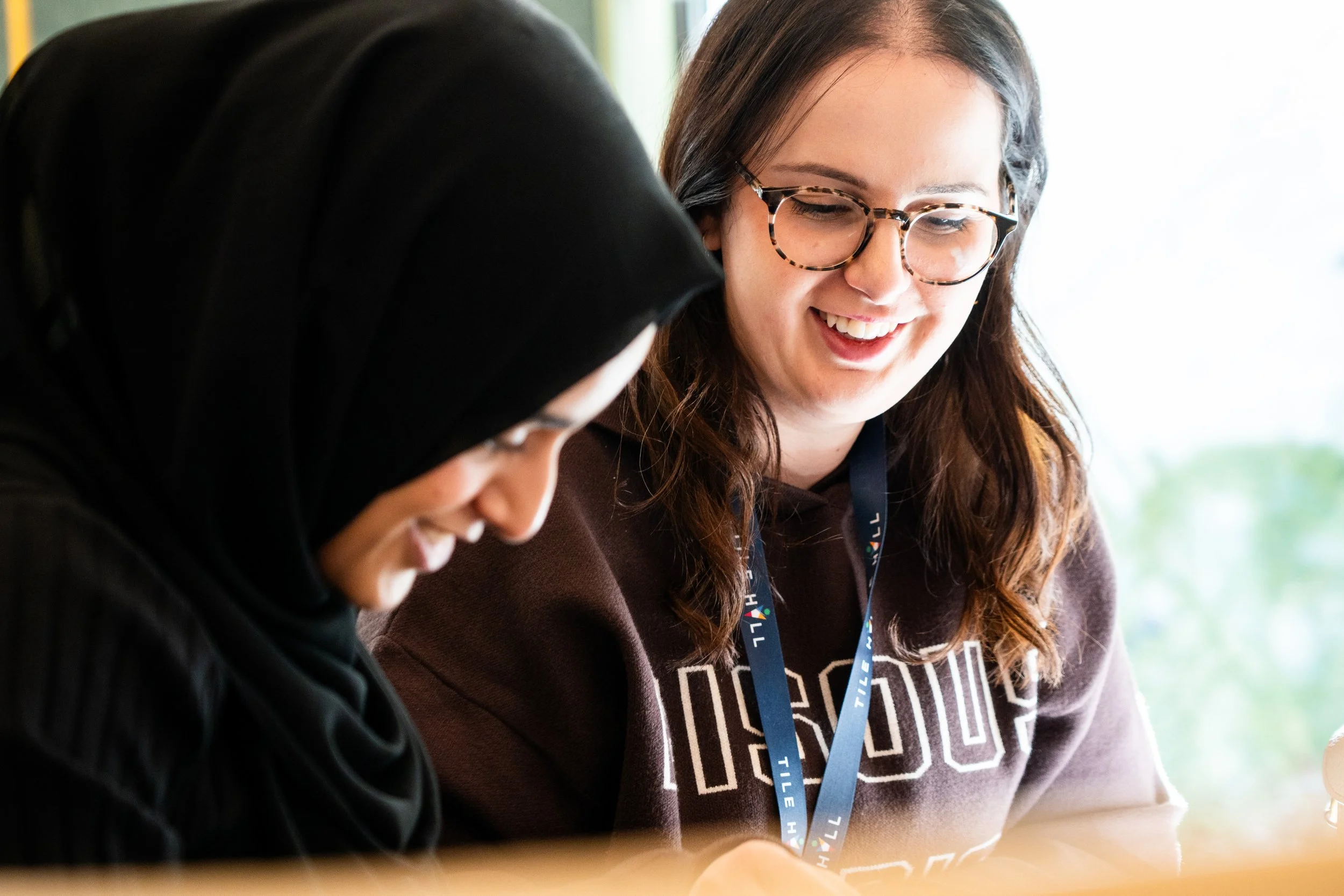 Two women with long hair and glasses smiling and engaged in a conversation, sitting at a table in a well-lit space.