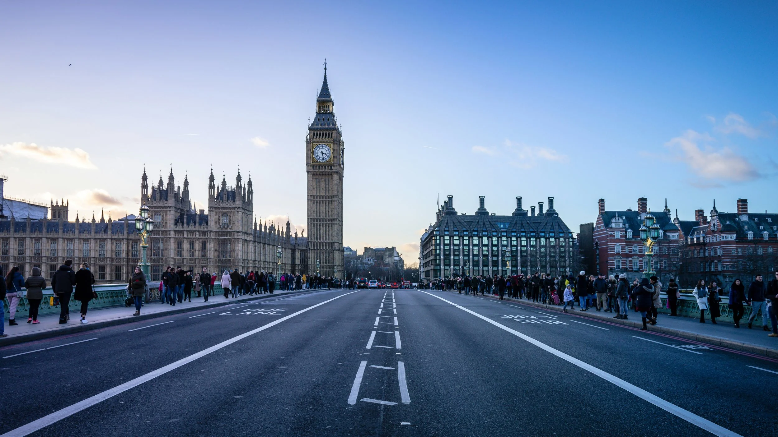 Big Ben clock tower and Palace of Westminster in London, viewed from across Westminster Bridge with crowds of people walking along the bridge, in the late afternoon or early evening.