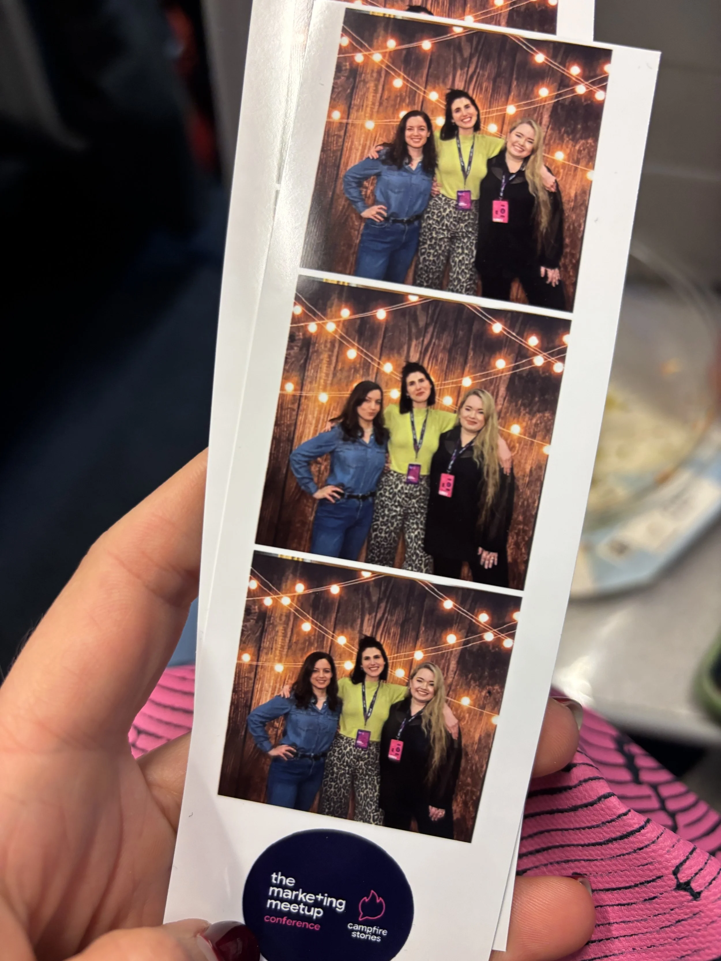 Photograph of three women at a conference with a wooden background and string lights, smiling and standing close together. The bottom of the photo has a logo for 'the marketing meetup conference' and 'campfire stories'.
