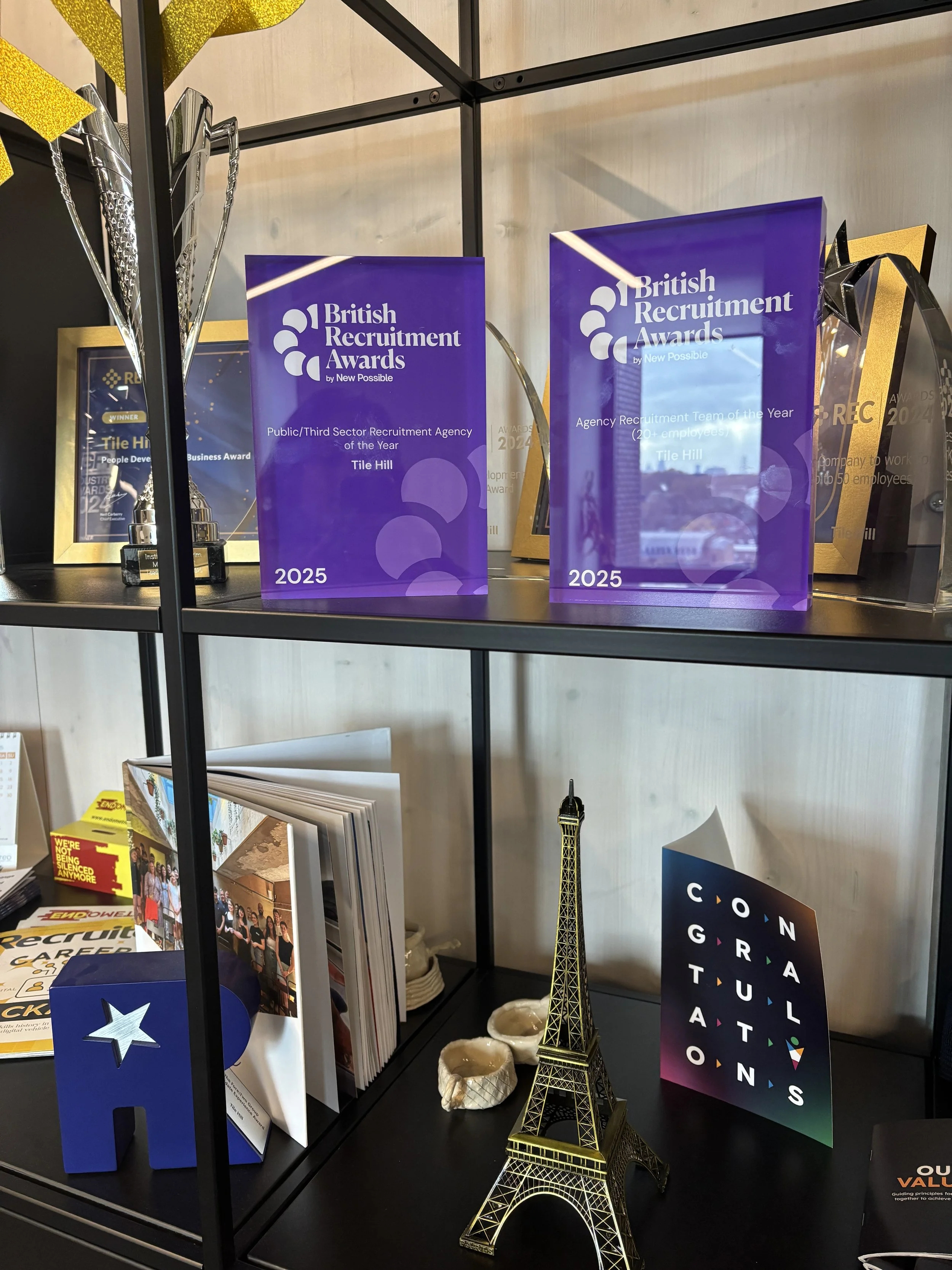 Shelving unit displaying awards, brochures, a miniature Eiffel Tower, and decorative items. The awards are purple with white text for the British Recruitment Awards 2025. The miniature Eiffel Tower is gold-colored and stands in front of the awards. There are also various brochures and framed certificates behind the awards.