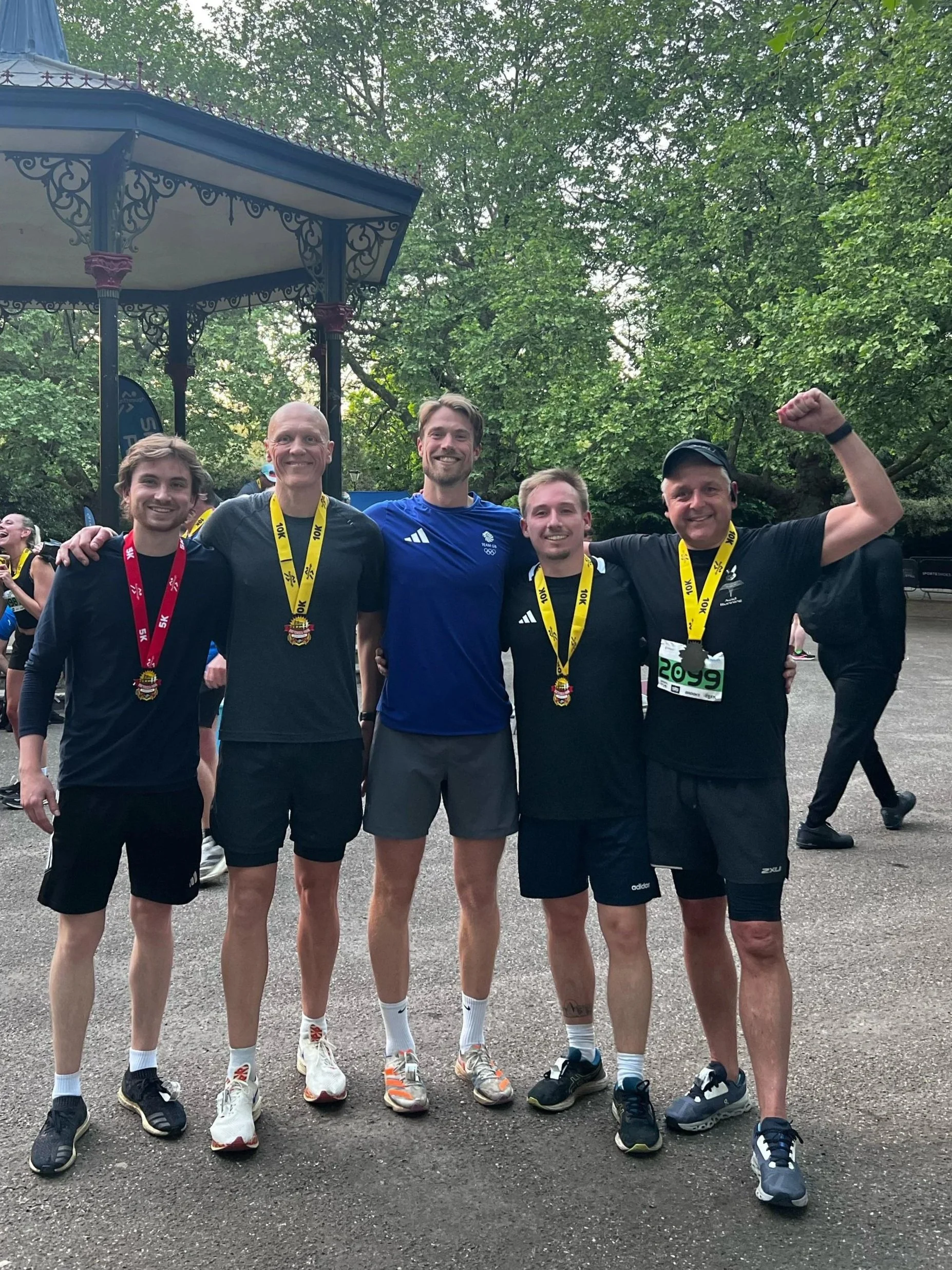 Five runners standing together outdoors, wearing athletic clothes and medals, smiling, after completing a race, with a park and some trees in the background.