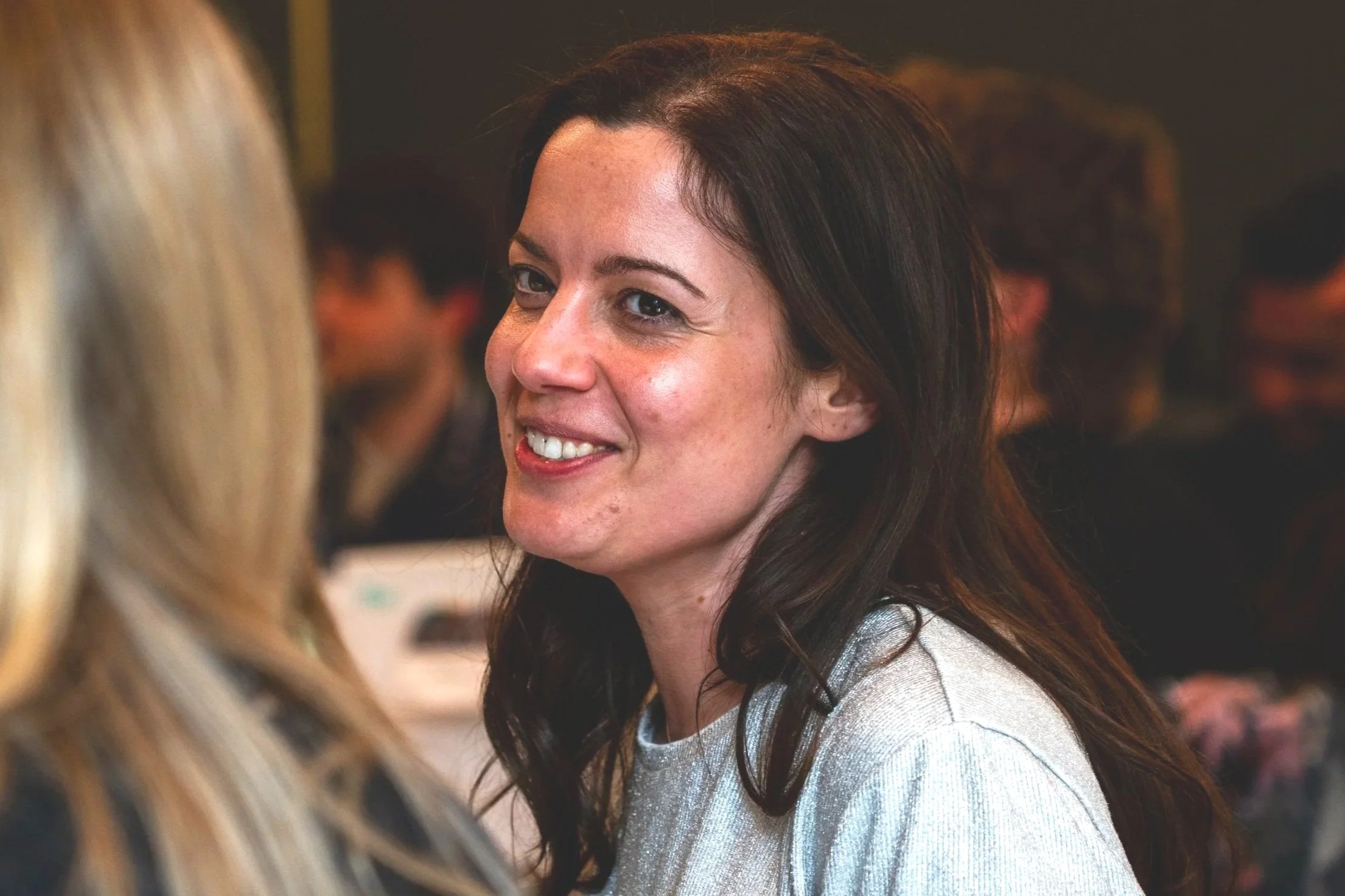 A woman with dark wavy hair smiling and talking to another woman with blonde hair in a crowded indoor setting.