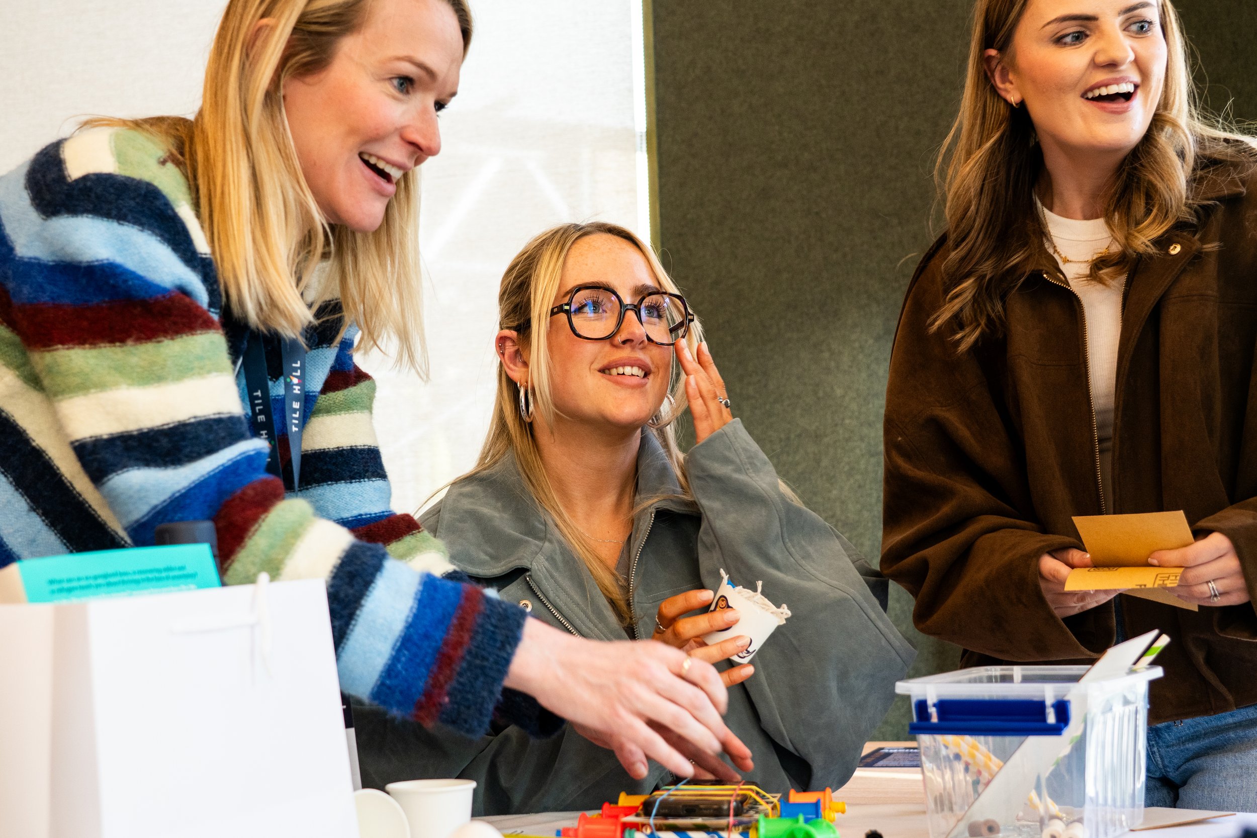 Three women at a table, smiling and engaging in an activity with colorful items and paper, in a casual setting.