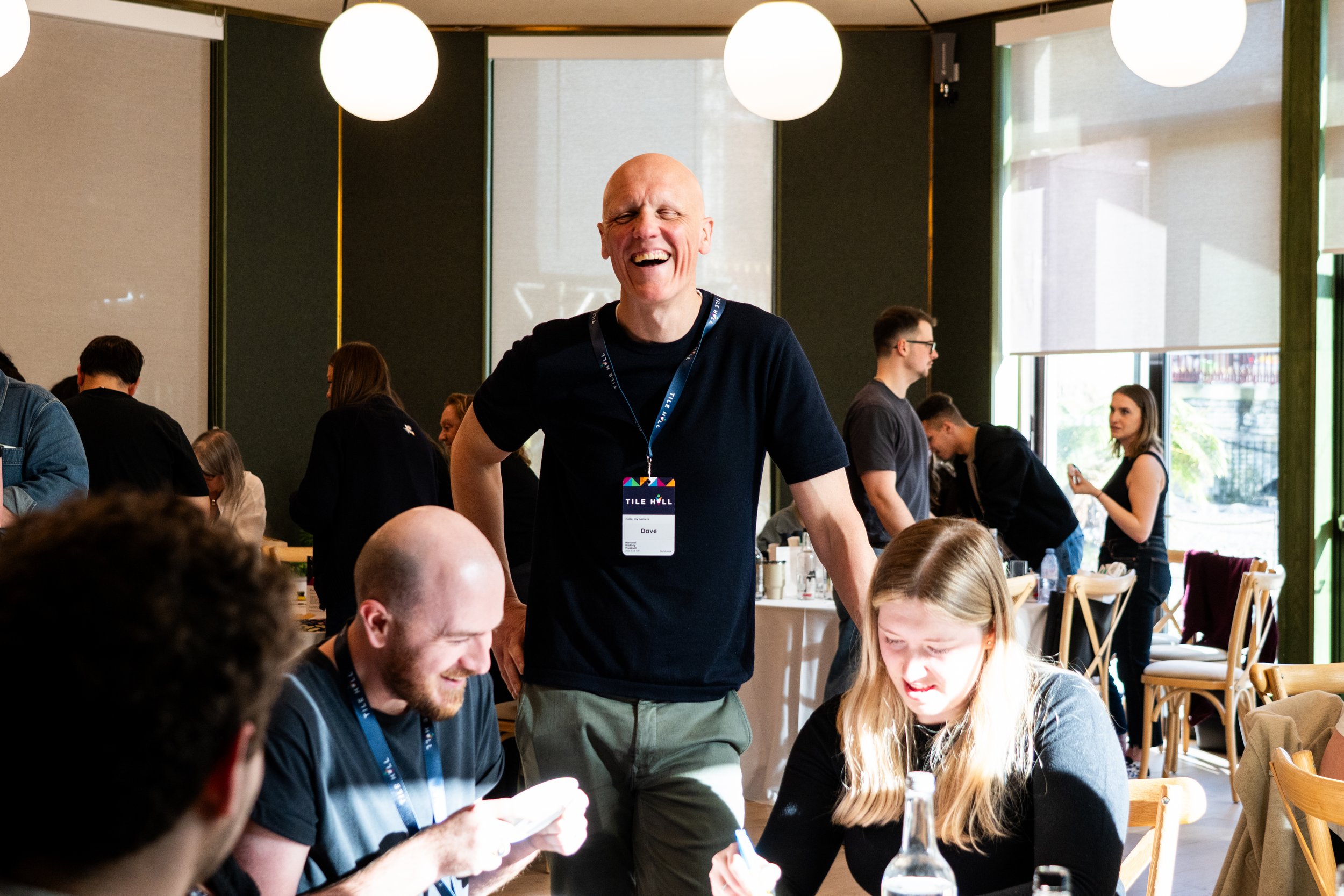A group of people in a bright, modern restaurant or conference room, with a man in the center smiling and wearing a name badge that reads 'Dave' at a conference or event titled 'Tile Hill.' Others are seated around tables, engaging with phones and each other.