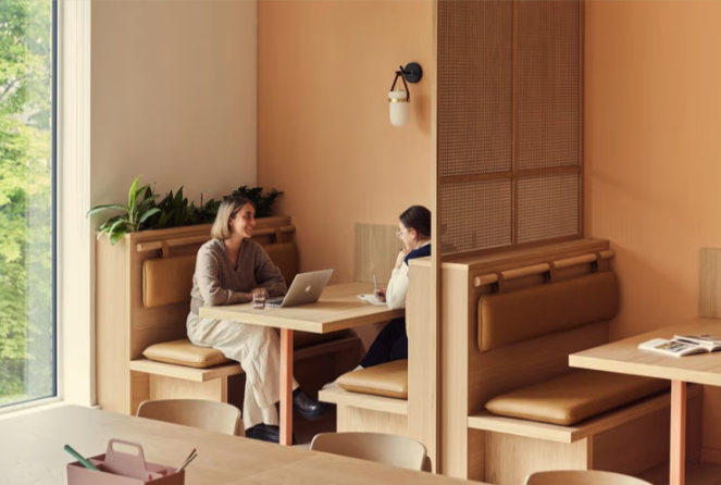 Two women sitting and talking at a booth in a cozy cafe with a beige wall, a wall sconce, and a large window with greenery outside.