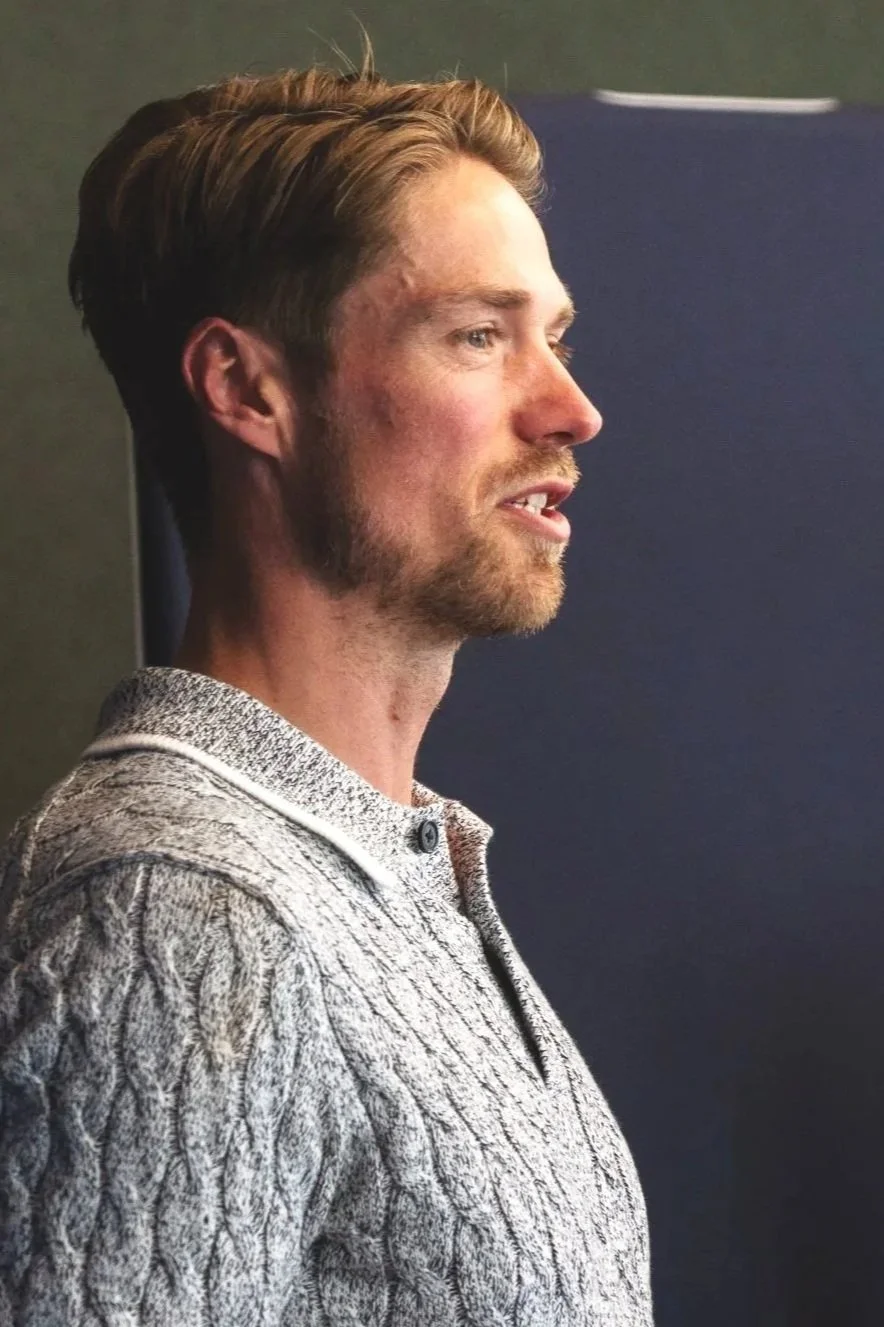 Profile of a young man with light brown hair, a beard, wearing a gray knit sweater, standing against a dark background.