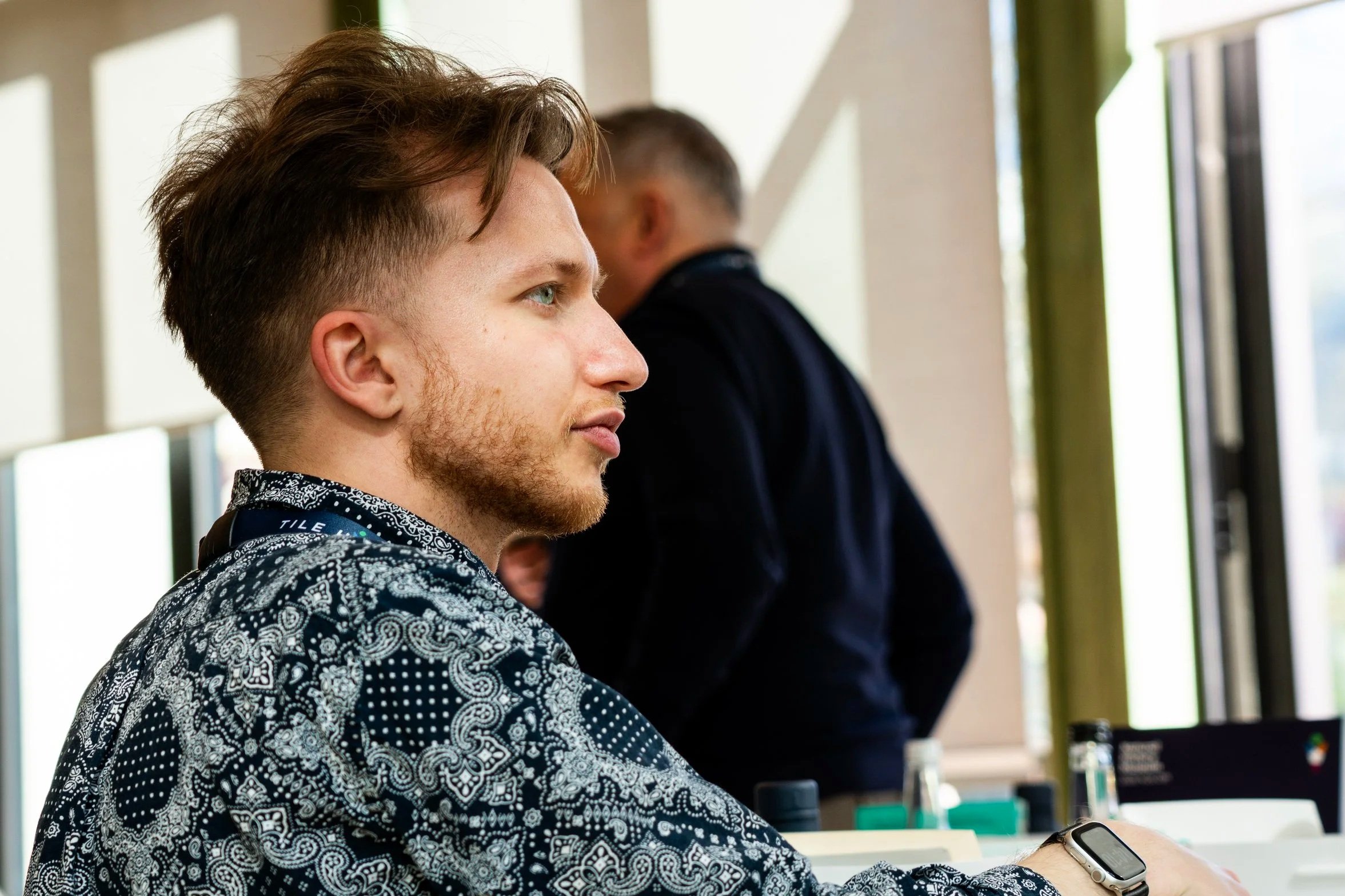 A young man with short, styled hair and a beard, wearing a black and white patterned shirt, sits at a table with a smartwatch on his wrist. He appears focused, possibly looking at a screen. In the background, another person with short, dark hair and a dark shirt is standing next to a window, with sunlight coming in.