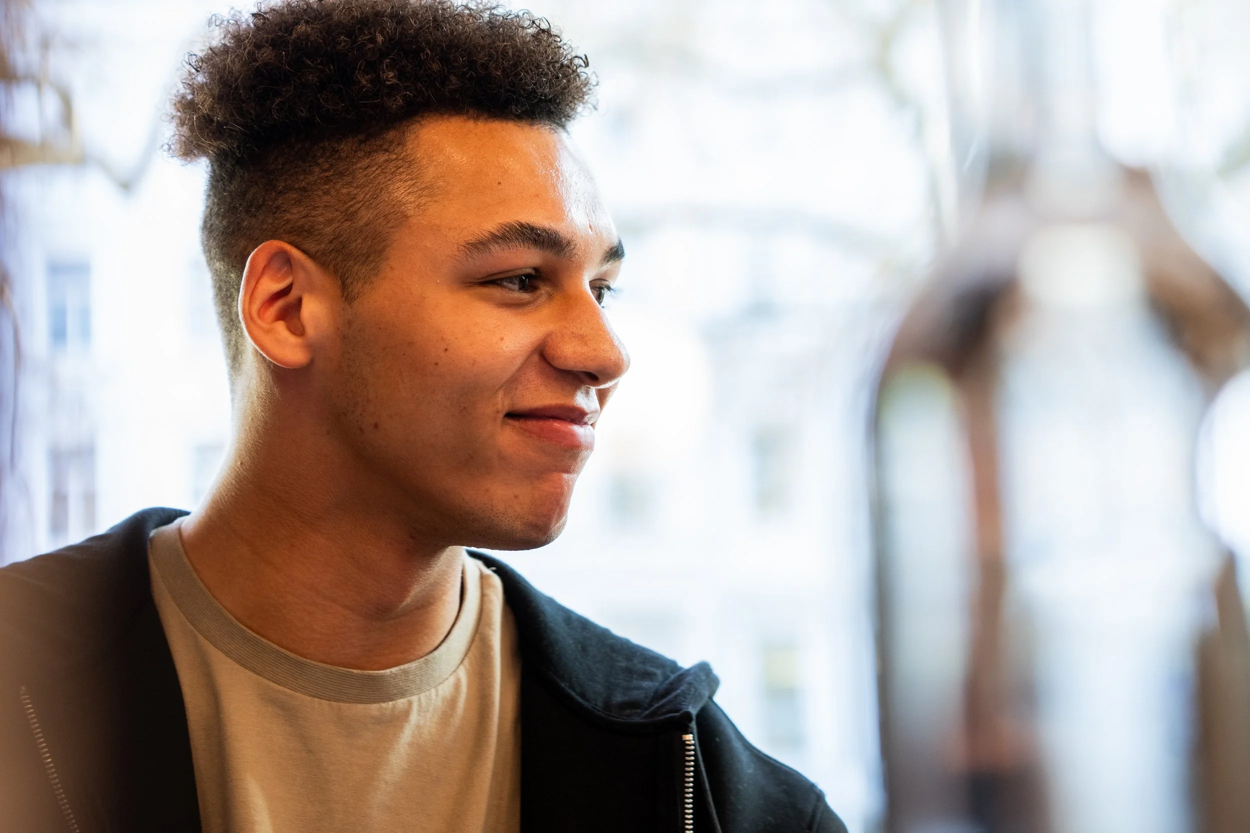 A young man with curly hair and a slight smile, wearing a beige t-shirt and a black jacket, standing indoors with a bright, blurred background.