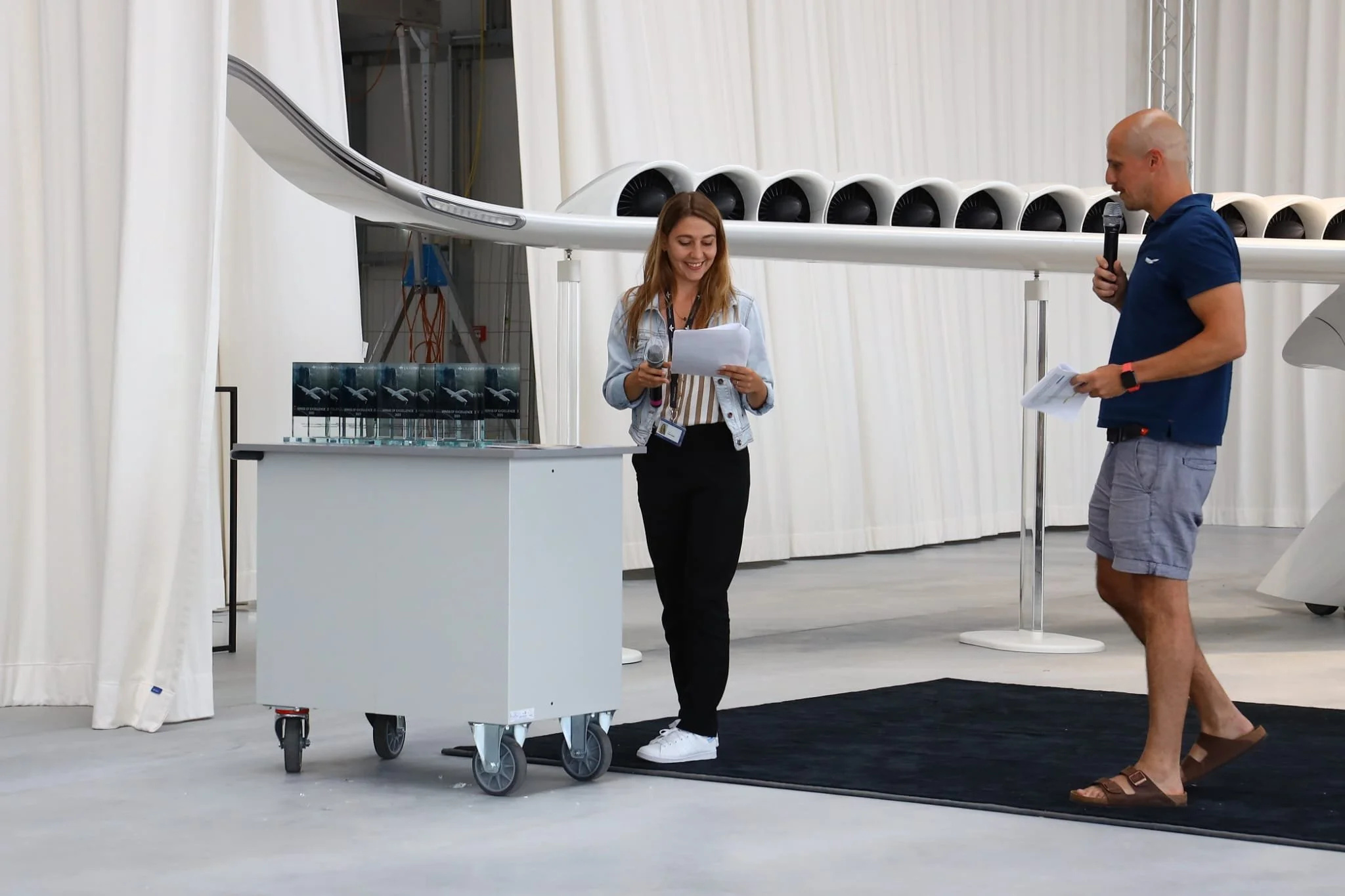 A woman holding a microphone and looking at papers stands. A man in shorts and a blue shirt with a microphone looks towards her. They are in a large, white, enclosed space with white curtains and technical equipment in the background.
