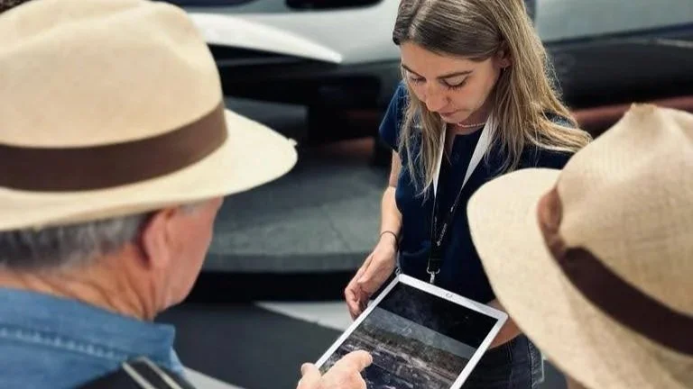 A woman showing a tablet to two elderly men wearing hats in a museum or gallery setting.