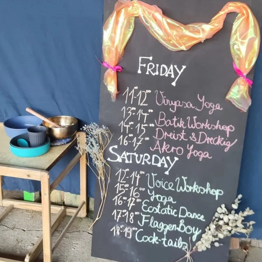 A chalkboard sign listing yoga, dance, and workshop classes scheduled for Friday and Saturday, decorated with pastel-colored fabric tied with pink ribbons, next to a small table with cups and a singing bowl, and dried flowers underneath.