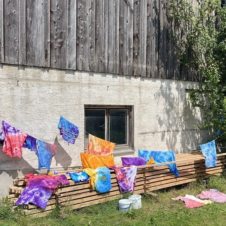 Colorful tie-dye clothing hanging on a clothesline outside a gray building with a wooden wall and a small window, with trees and grass nearby.