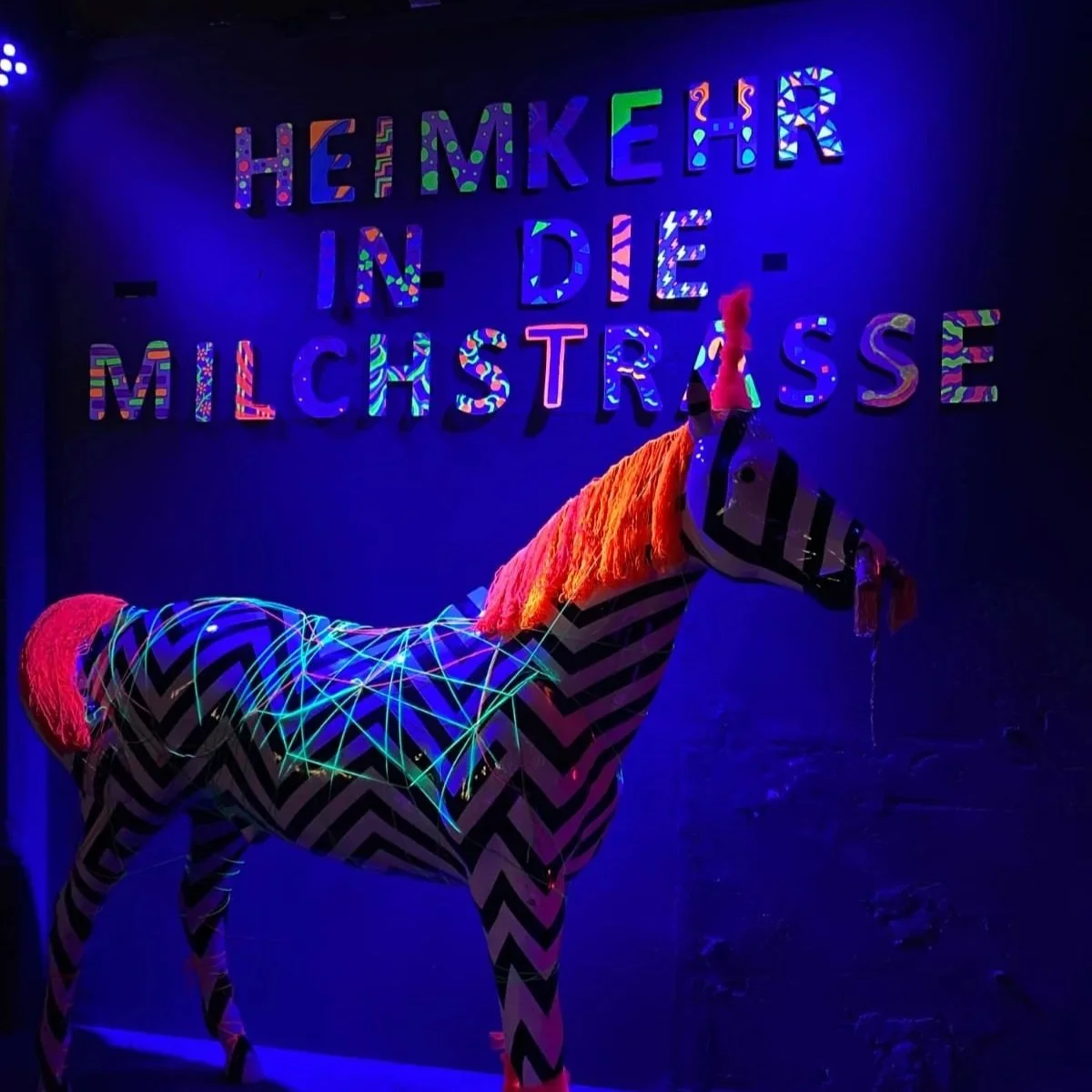 A zebra sculpture with black and white stripes, neon lights on its body, and orange and pink accents on its mane and tail, standing in front of a colorful sign that says 'THE MAKER IN THE MICHTHRASE' against a dark background.