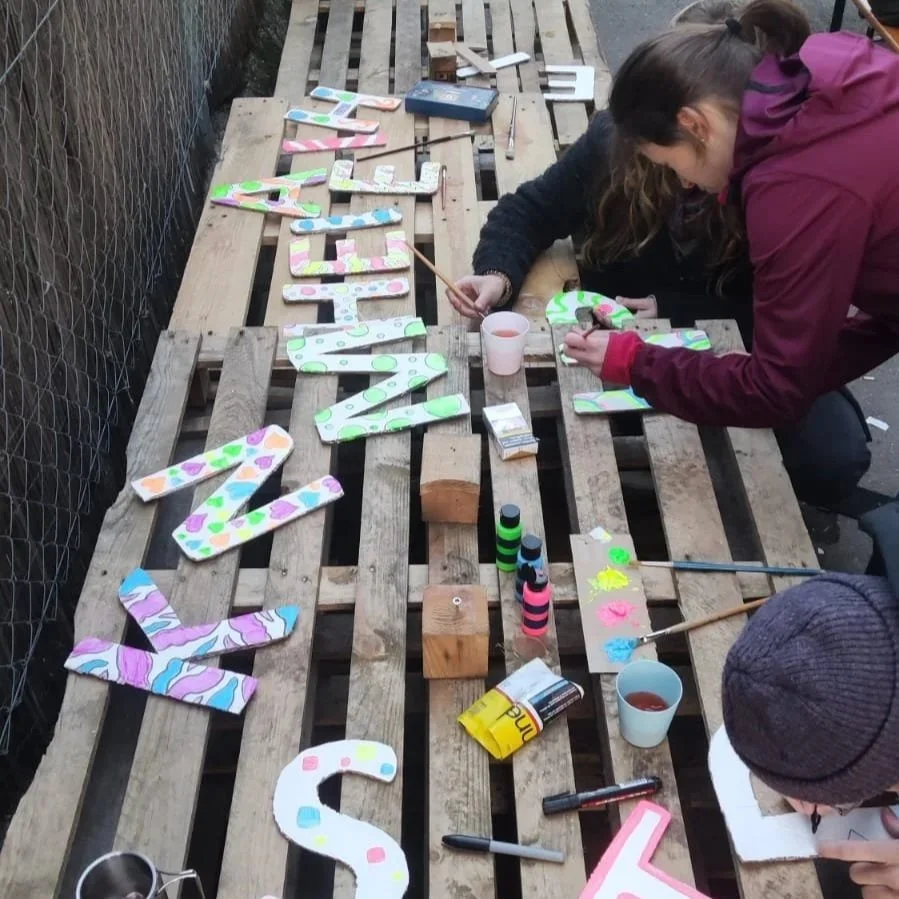People painting colorful letter decorations on a wooden pallet table outside.