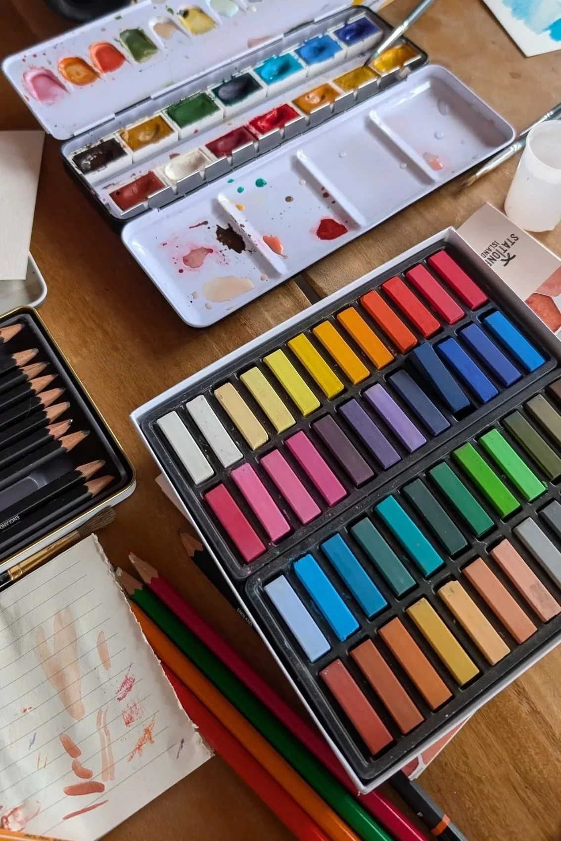 Overhead view of a watercolor art setup including paint palettes with various colors, a box of pastel chalks organized by color, colored pencils, and a sketchbook with scribbles and smudges.