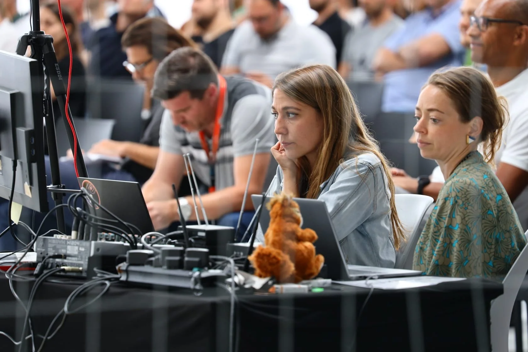 People sitting at a conference or seminar, focused on a presentation or screen, with electronics and cables on the table, and a stuffed teddy bear in front of a woman with long hair wearing a denim jacket.