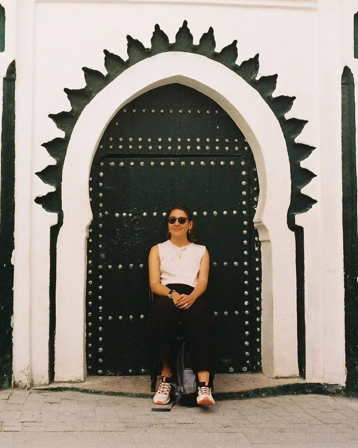 A woman in sunglasses and casual clothing sitting in front of an ornate arched black door with silver studs and decorative black sunburst accents.